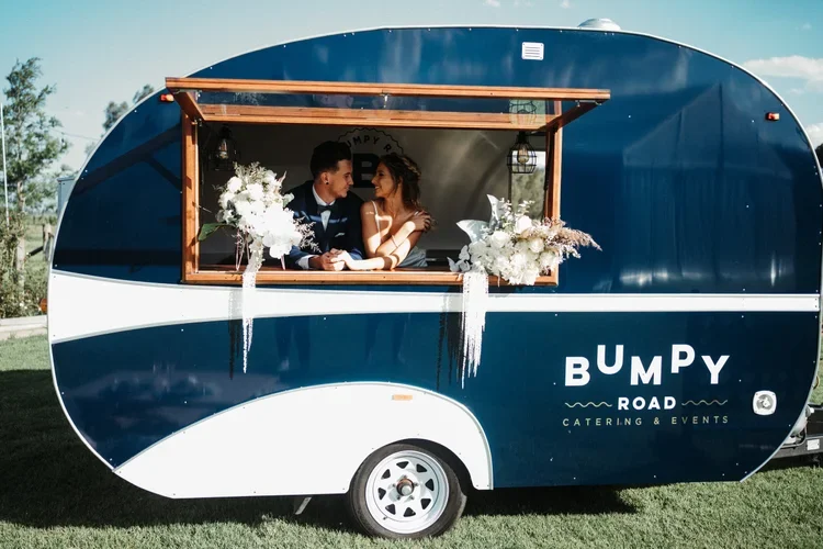 A couple dressed in wedding attire sitting inside a blue and white mobile catering trailer with floral decorations, celebrating their wedding.