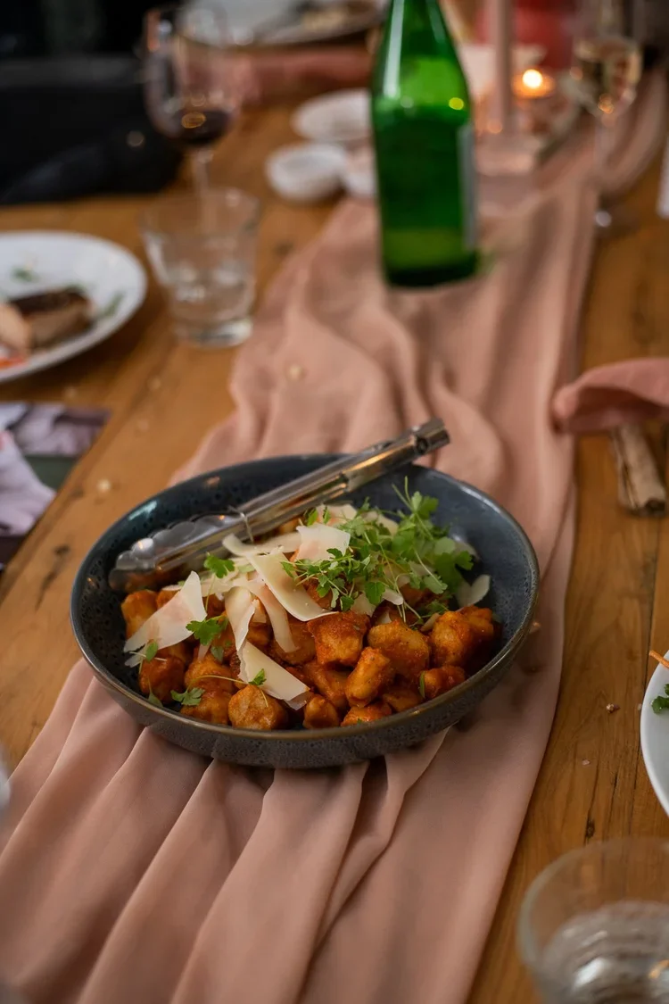 A black bowl of gnocchi topped with shaved Parmesan cheese, fresh herbs, and a pair of tongs, on a table with a pink tablecloth, surrounded by glasses and plates, in a cozy dining setting.