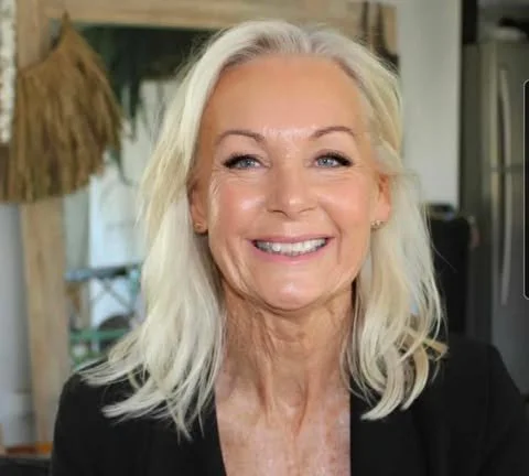 Smiling mature woman with long silver hair, wearing a black top, indoor setting.