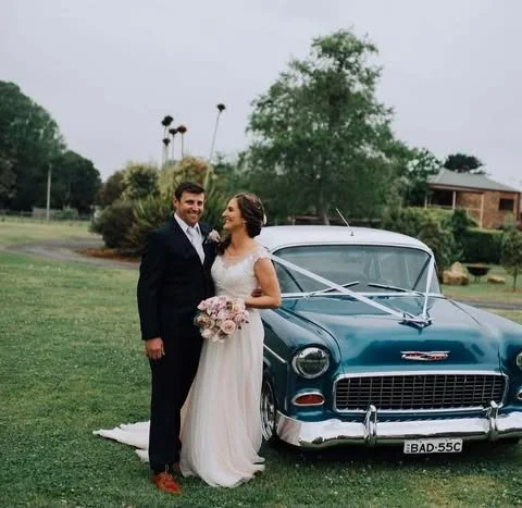 Bride and groom smiling and standing next to a decorated vintage blue car with a wedding license plate, on a grassy area with trees and a house in the background.