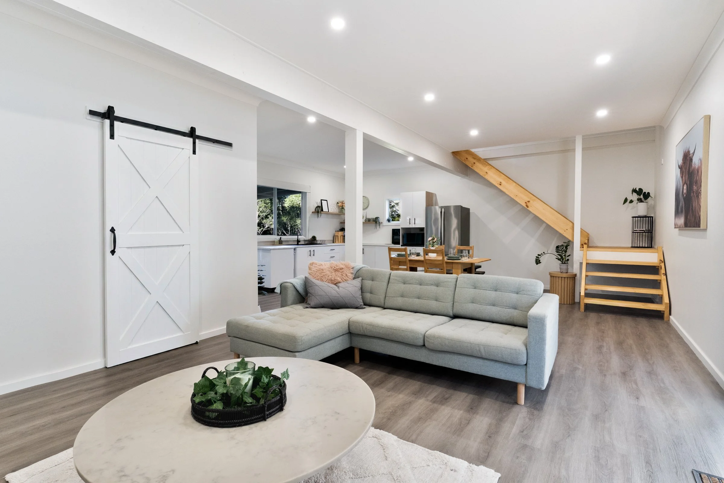 Open-concept living room with gray sectional sofa, round marble coffee table with greenery, white walls, wood flooring, and bright recessed lighting. Visible kitchen area in the background with white cabinets and stainless steel appliances. Wooden st