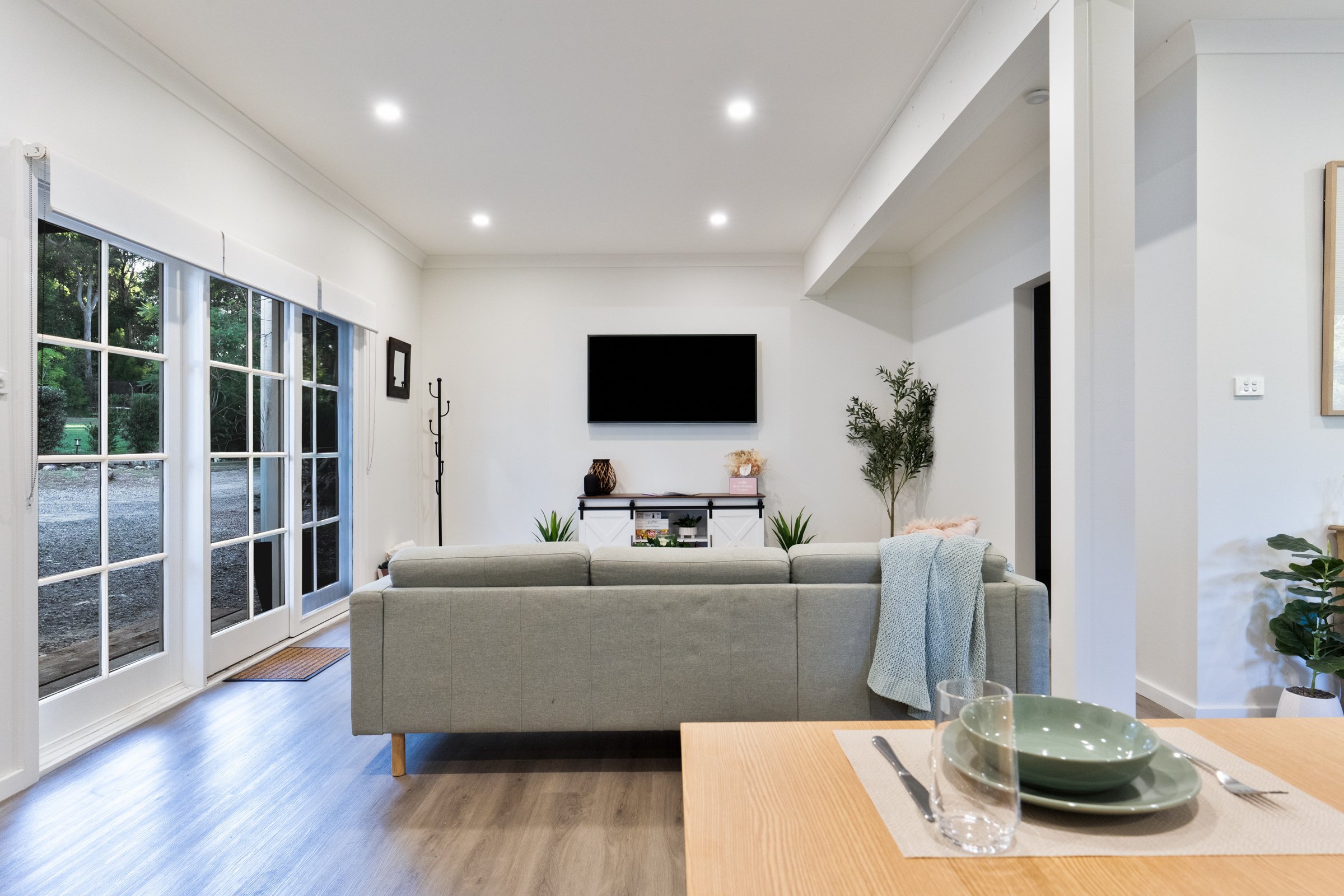 A bright, modern living room with white walls and ceiling, a wall-mounted flat-screen TV, a beige couch, large glass doors with white frames and views of greenery outside, and hardwood floors.