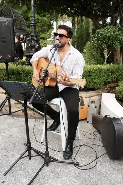 A man wearing sunglasses and a white shirt is playing an acoustic guitar and singing into a microphone outdoors. There are musical equipment, a music stand, and trees in the background.