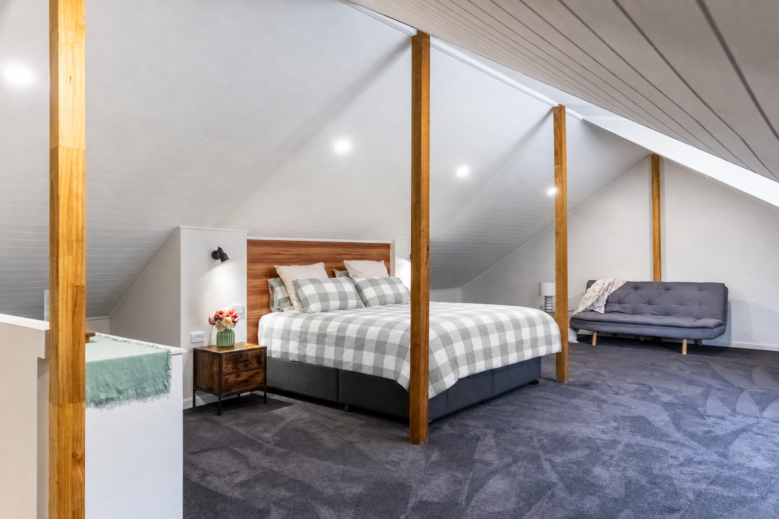 Bedroom with a sloped ceiling, a large bed with plaid bedding, a wooden headboard, a side table with a vase of flowers, a gray sofa, and a lamp, carpeted floor, and wooden support beams.