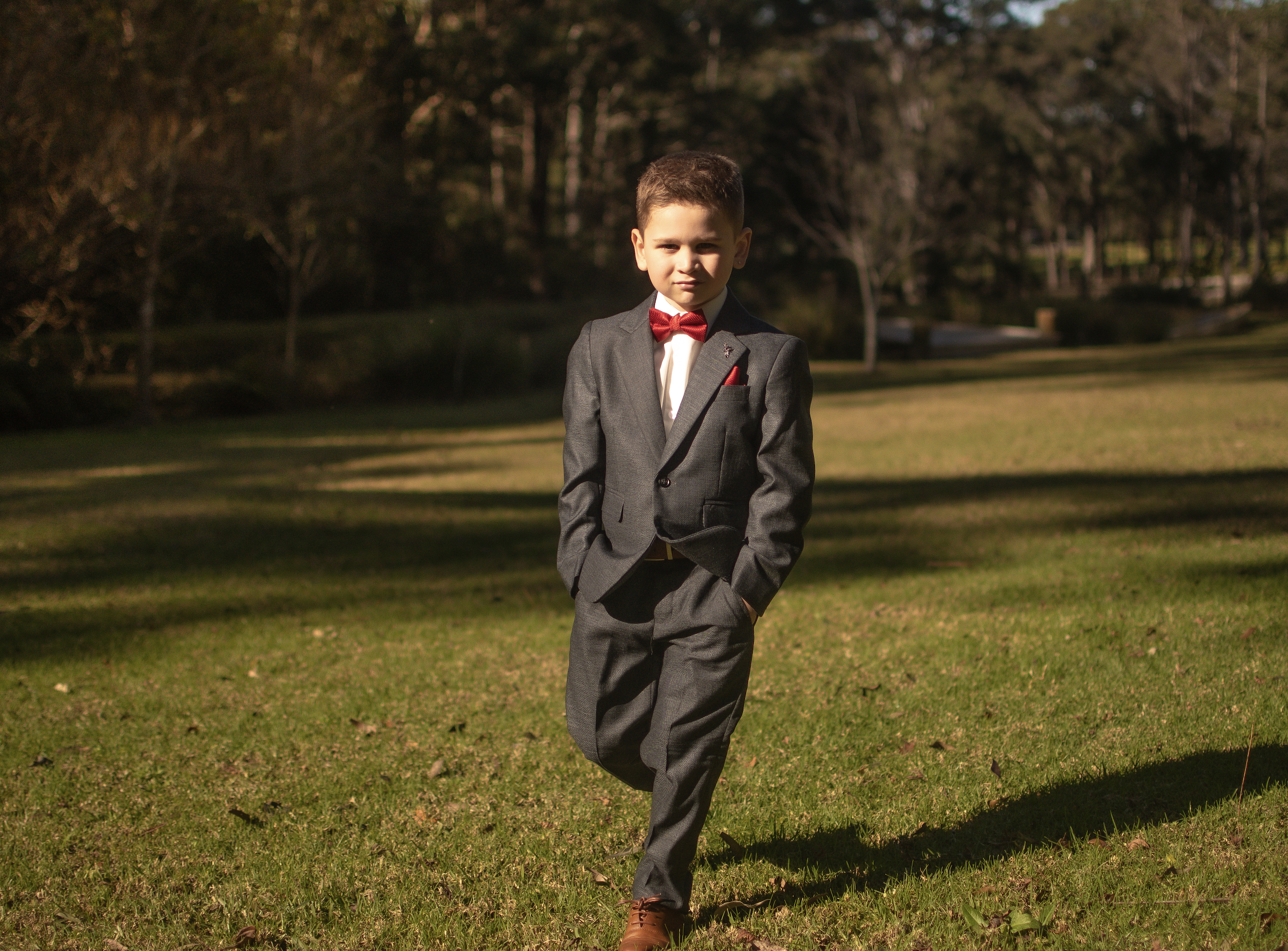 A young boy dressed in a formal suit with a red bow tie and pocket square, standing outdoors on a grassy field with trees in the background.