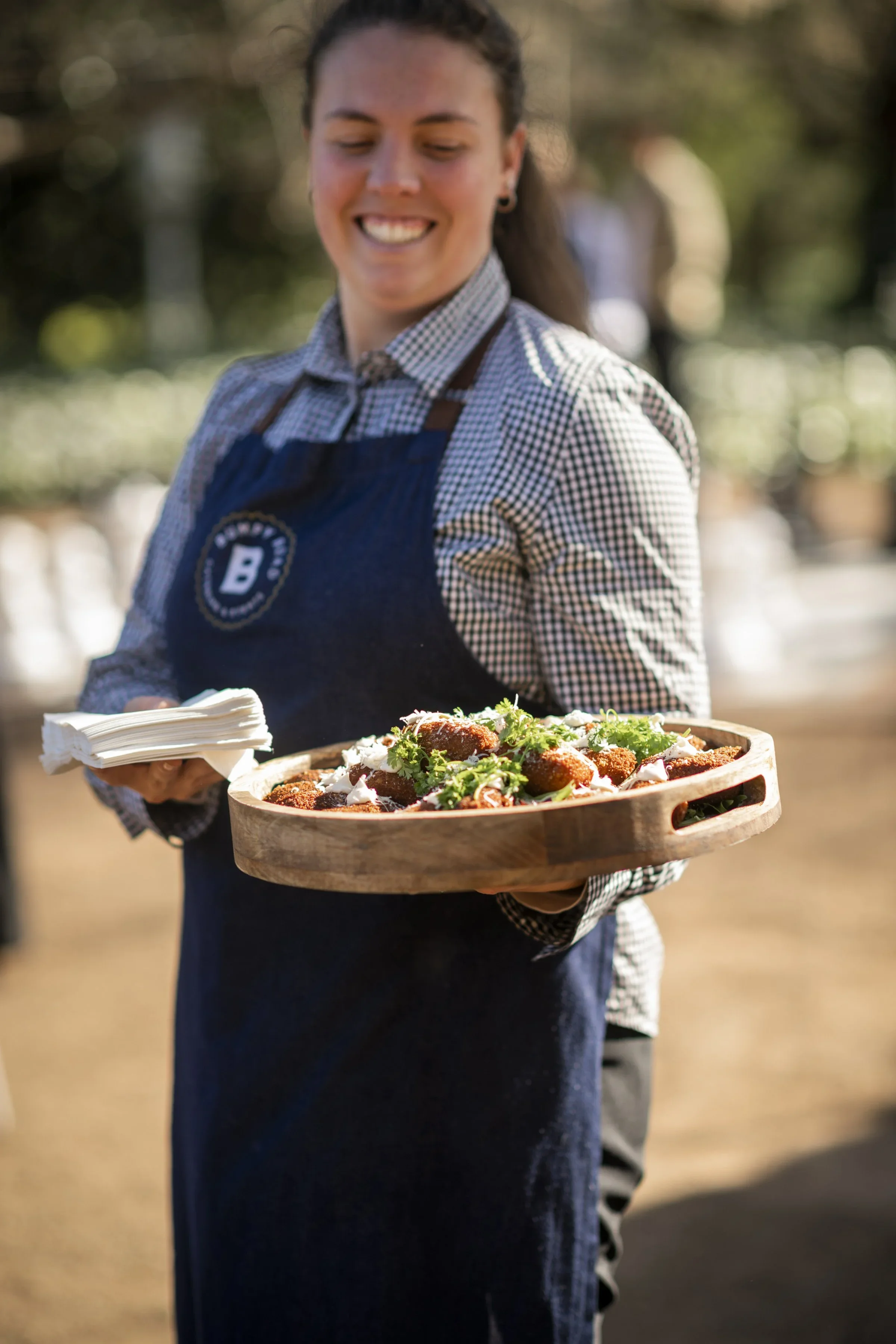 A woman wearing a checkered shirt and a denim apron with a logo, smiling while holding a wooden tray with fried food and garnished with herbs, outdoors in a sunny setting.