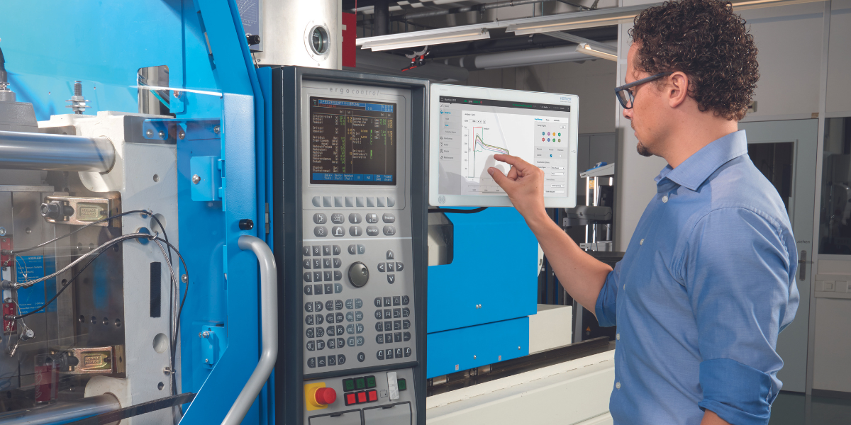 A man in a blue shirt and glasses is working with industrial machinery, pointing at a computer screen showing data and graphs in a manufacturing or laboratory setting.