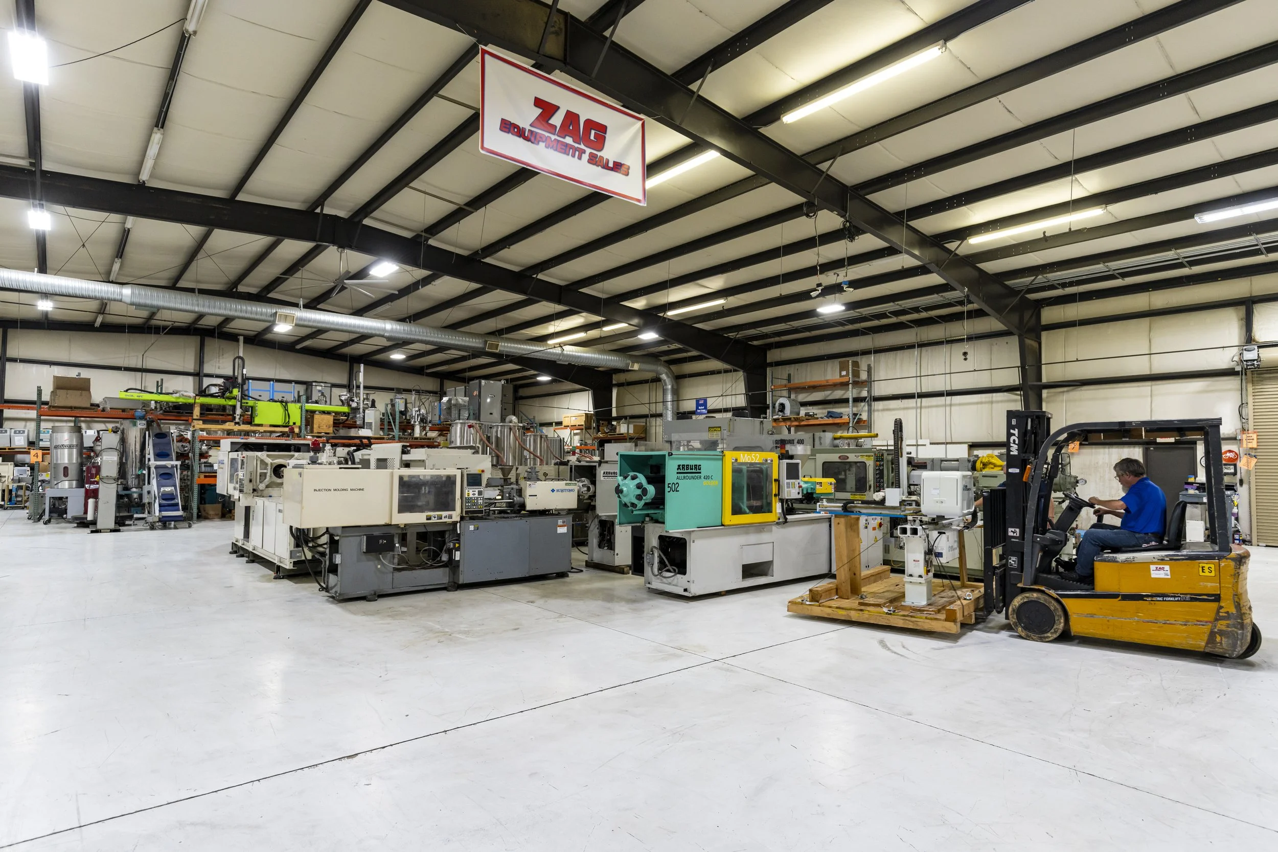 A spacious industrial warehouse with various manufacturing machines and equipment, including a worker operating a forklift. A hanging sign reads 'ZAG Equipment Sales'.