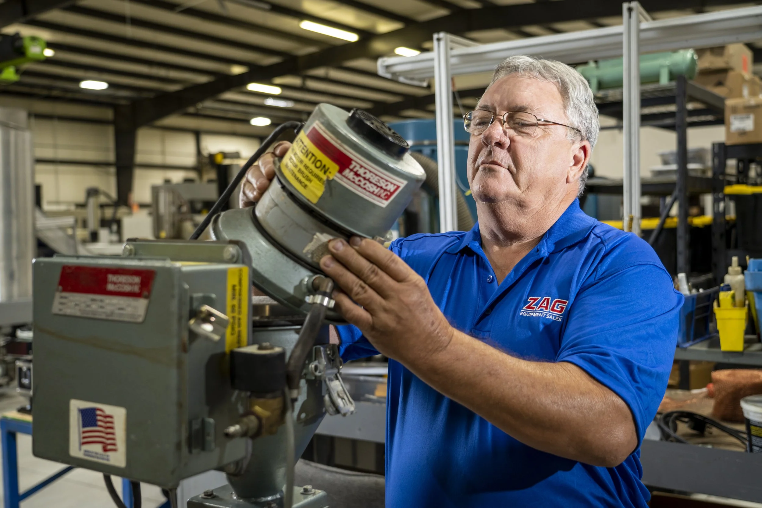 A man in a blue shirt with the logo ZAG Equipment Sales working with industrial machinery in a workshop or factory setting.