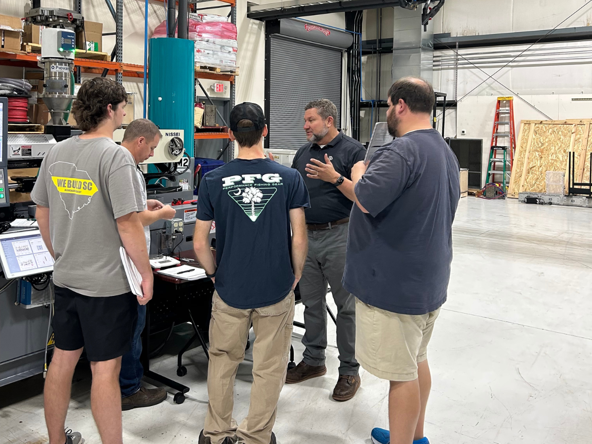 Five men standing in a workshop, engaged in conversation around a workbench with tools and equipment.