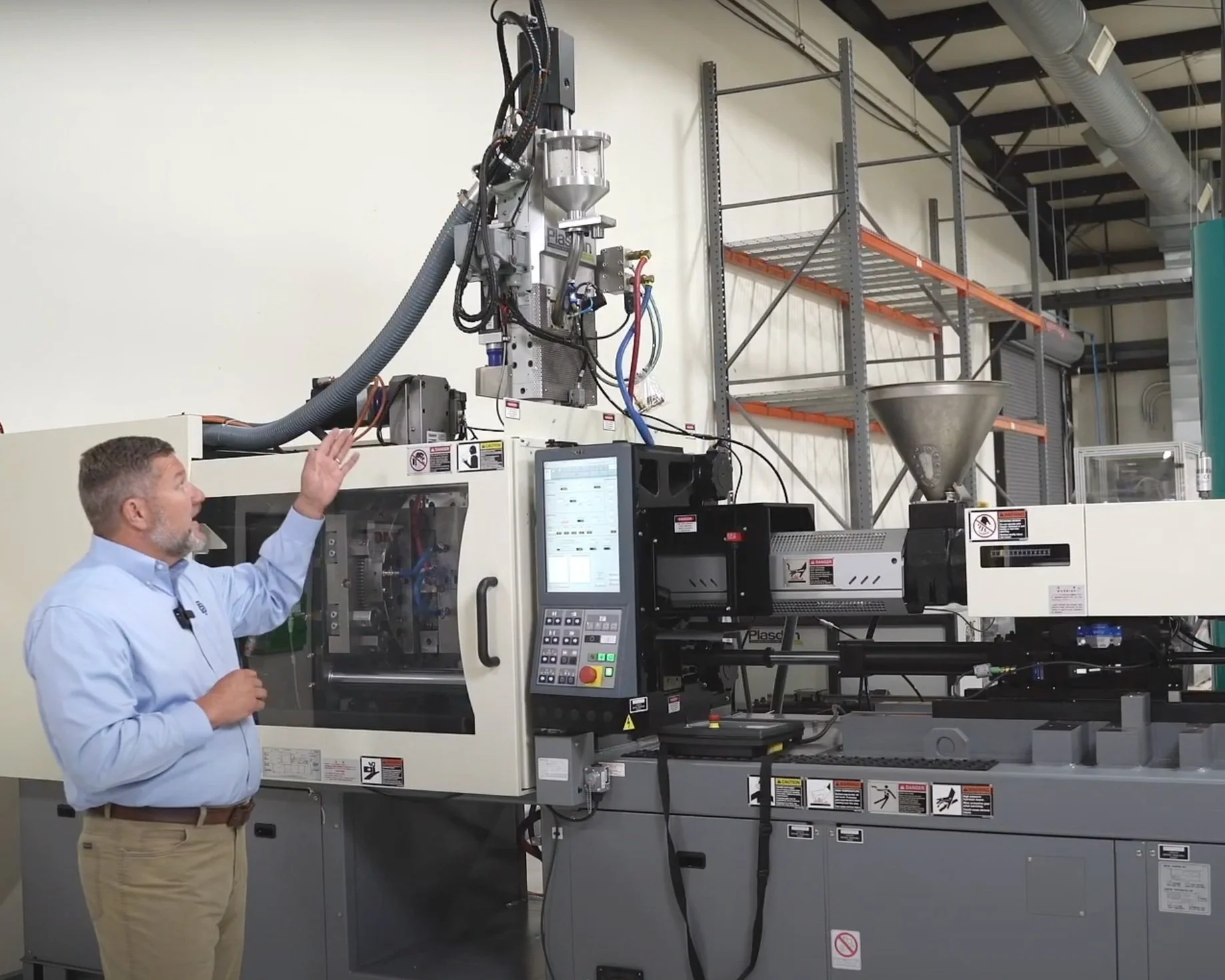 A man in a light blue shirt and khaki pants gesturing in front of a large industrial injection molding machine in a warehouse or factory setting.