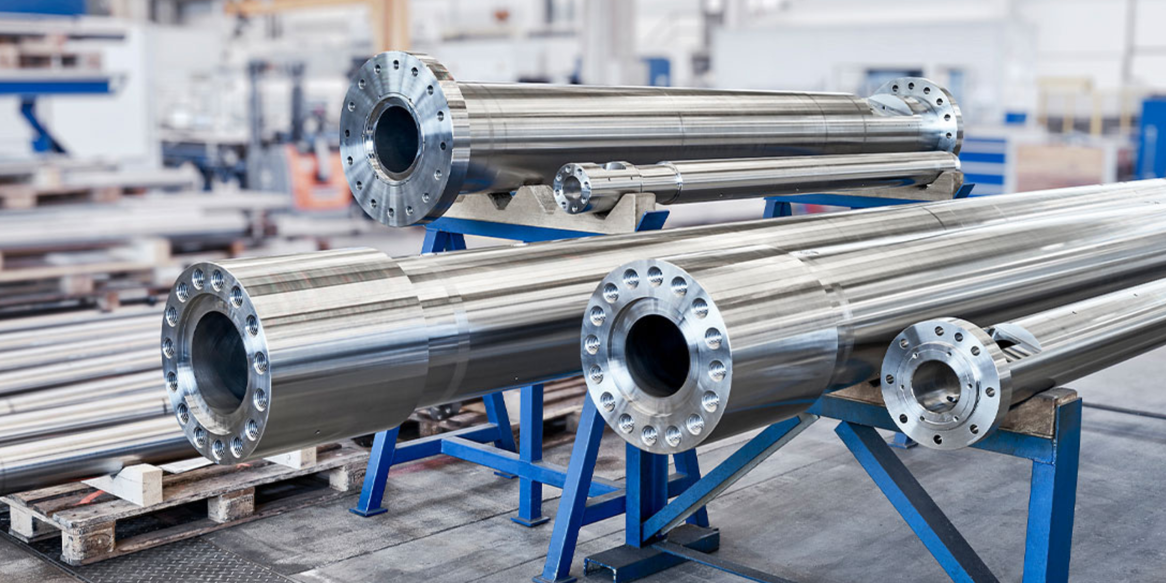 Large polished metal industrial cylinders with flanged ends on blue metal supports in a manufacturing facility.