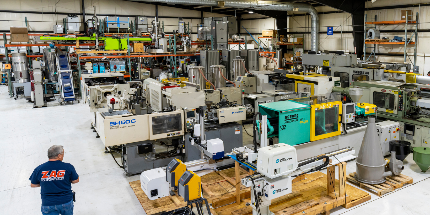 A man wearing a blue ZAG Equipment Sales shirt observes industrial manufacturing machines inside a large warehouse. Multiple CNC and injection molding machines are visible, with tools, pallets, and shelves in the background.