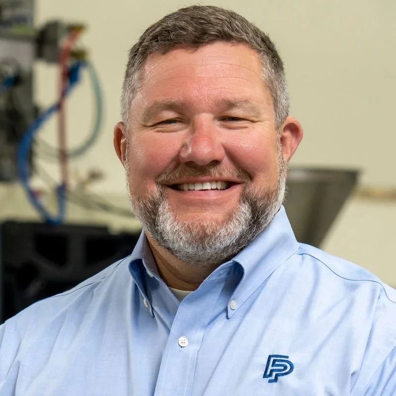 A smiling man with short gray hair and a beard, wearing a light blue button-up shirt with an embroidered logo on the chest, standing in an office or laboratory with equipment and wires in the background.