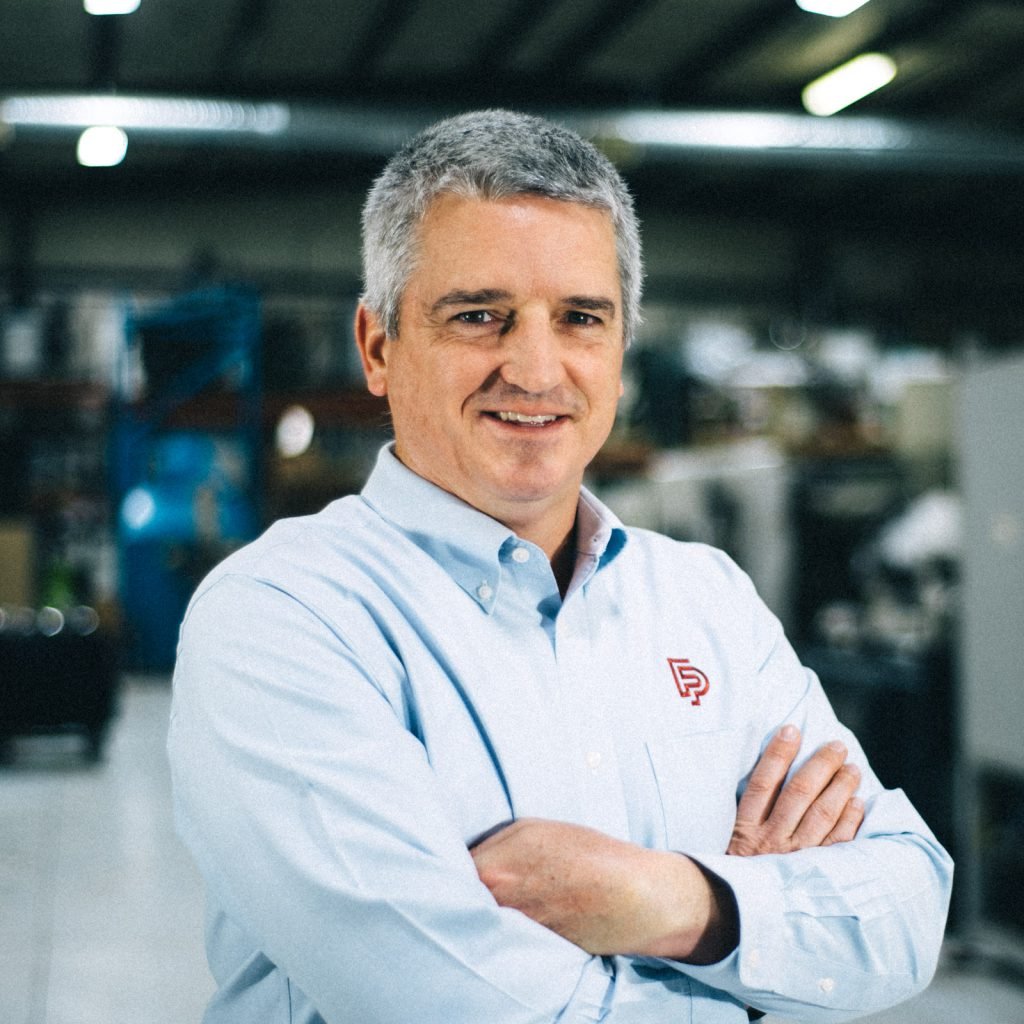A man with short gray hair wearing a light blue button-up shirt with a logo, standing with arms crossed in an industrial or warehouse setting.