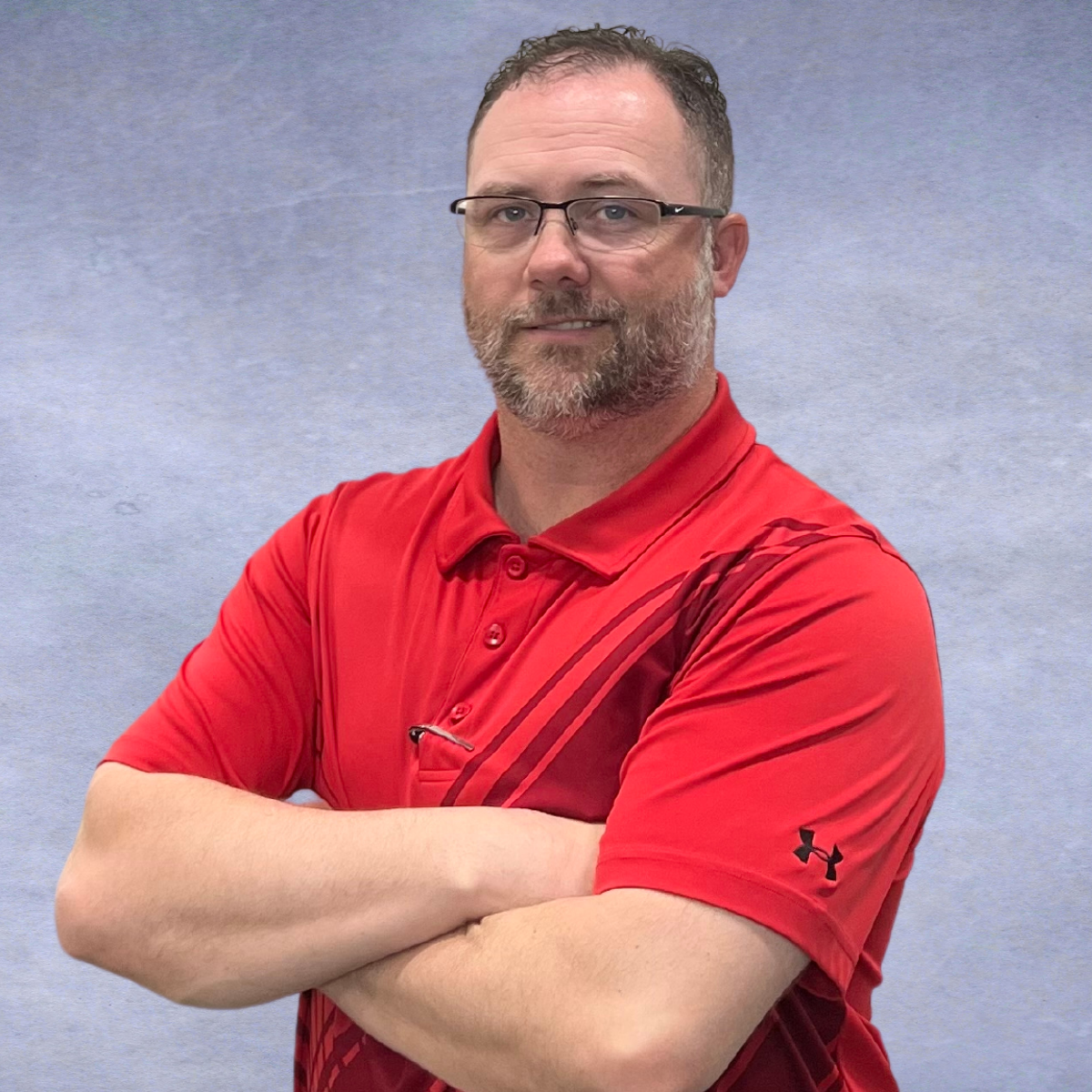 A man with glasses and a beard, wearing a red Under Armour polo shirt, standing with his arms crossed against a plain gray background.