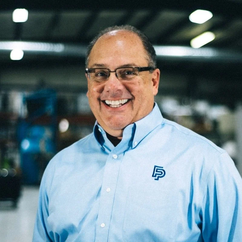 Smiling man with glasses wearing a light blue collared shirt, logo on chest, indoors with blurred background.