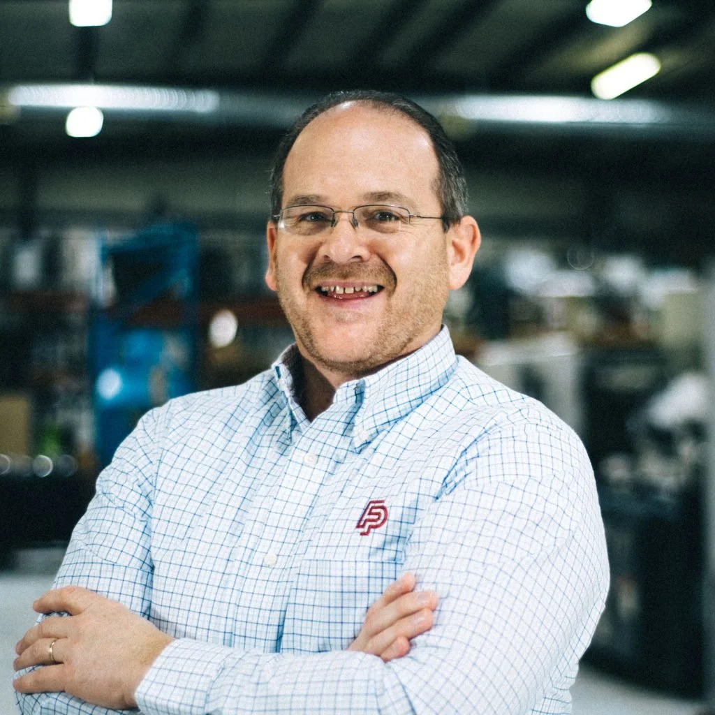 A man with glasses and a checkered shirt standing with arms crossed, smiling in an industrial or warehouse setting.