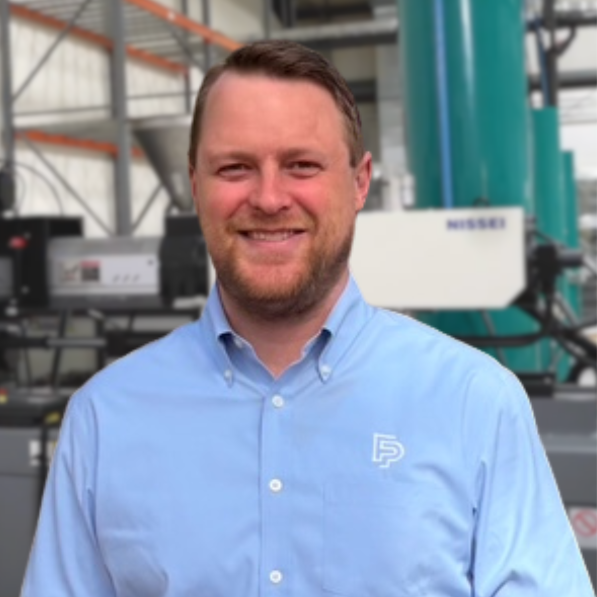 A smiling man with short brown hair and a beard wearing a light blue collared shirt with a logo, standing in an industrial or manufacturing setting with machinery and equipment in the background.