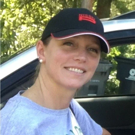 Woman smiling outdoors, wearing a black and red cap, pearl earrings, and a light purple shirt