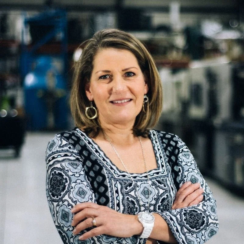 A woman with shoulder-length brown hair smiling with arms crossed, wearing a patterned black and white top, earrings, a watch, and a necklace, standing in a workshop or industrial setting with blurred machinery in the background.