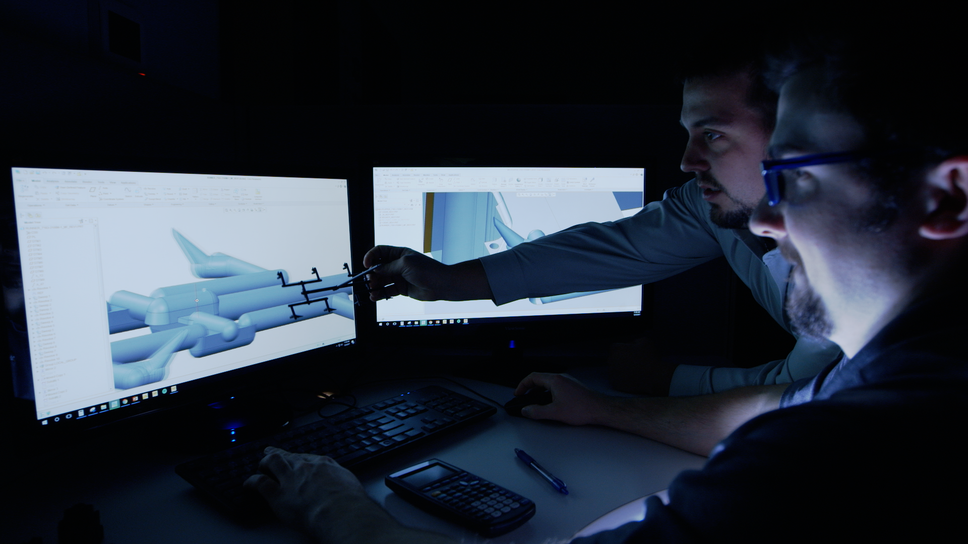 Two engineers working on a computer with 3D pipe design on dual monitors in a dark room.