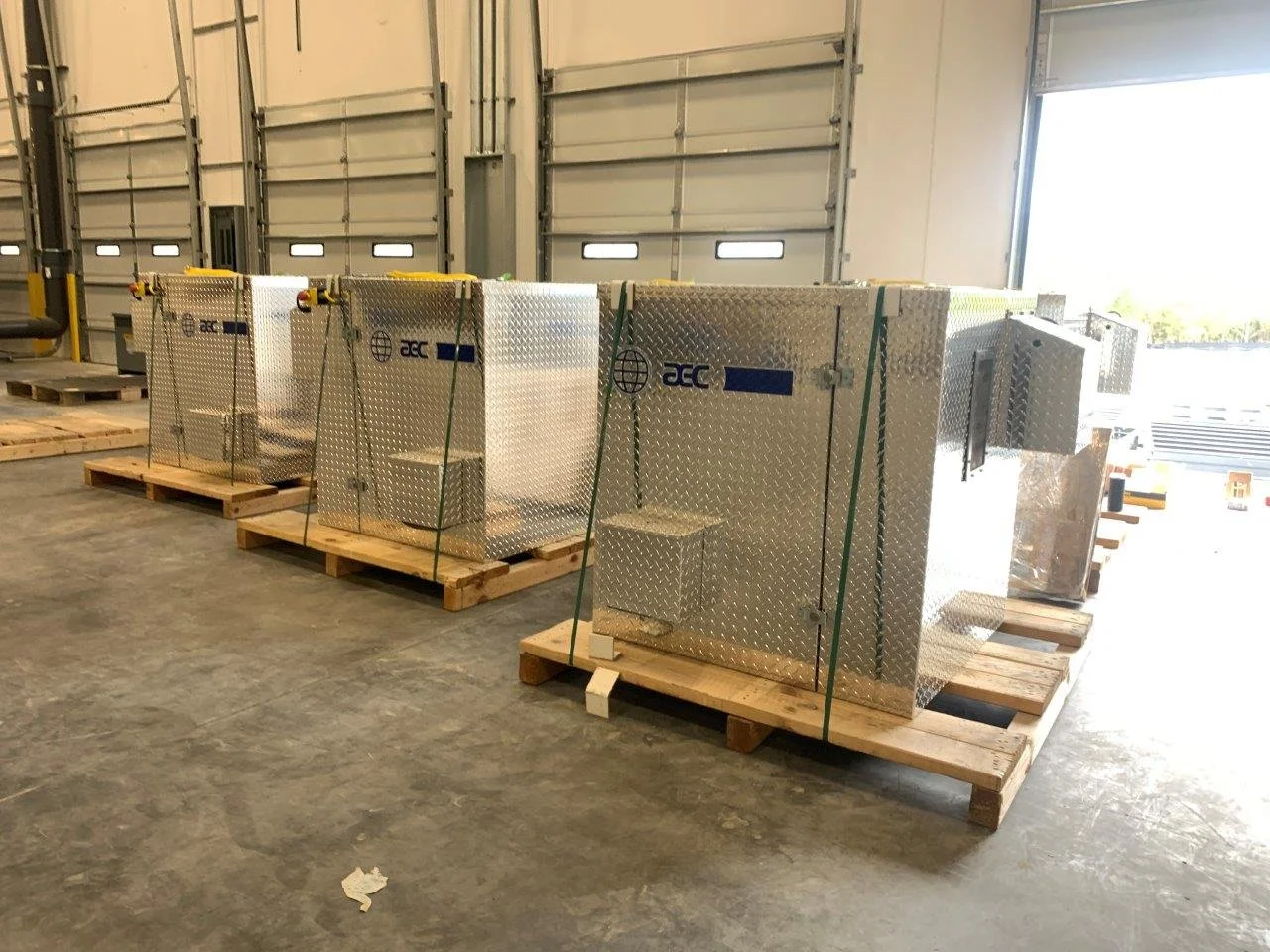 Four metallic industrial boxes on wooden pallets inside a warehouse, with a large open garage door in the background.