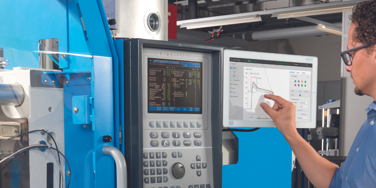 A man with curly hair and glasses is pointing at a computer screen showing a graph in an industrial or laboratory setting. Next to him are control panels and machinery, with exposed pipes and equipment in the background.