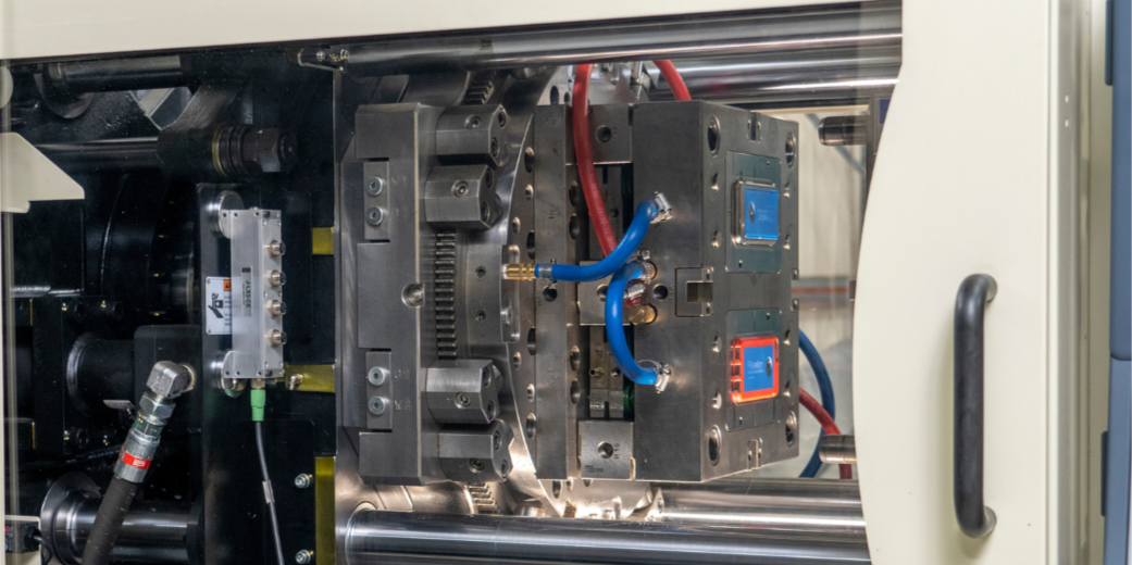 Close-up view of a precision industrial machine or injection molding equipment with metal components, hydraulic tubes, and control units inside a manufacturing environment.