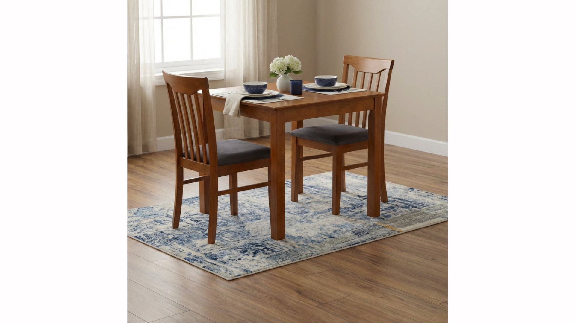 Dining table set for two with navy bowls, white plates, napkins, a vase of white flowers, and a candle in a blue holder, on a patterned rug in a room with wooden flooring and a window with sheer curtains.