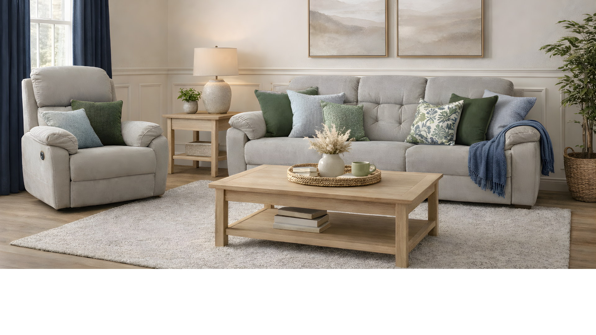 A cozy living room with a light gray recliner and a matching sofa, decorated with various green and patterned throw pillows. There is a wooden coffee table with a vase of dried flowers and a tray with a mug, placed on a textured cream rug. A side table with a lamp and a small potted plant, along with a large potted plant and window with dark blue curtains, complete the room's decor.