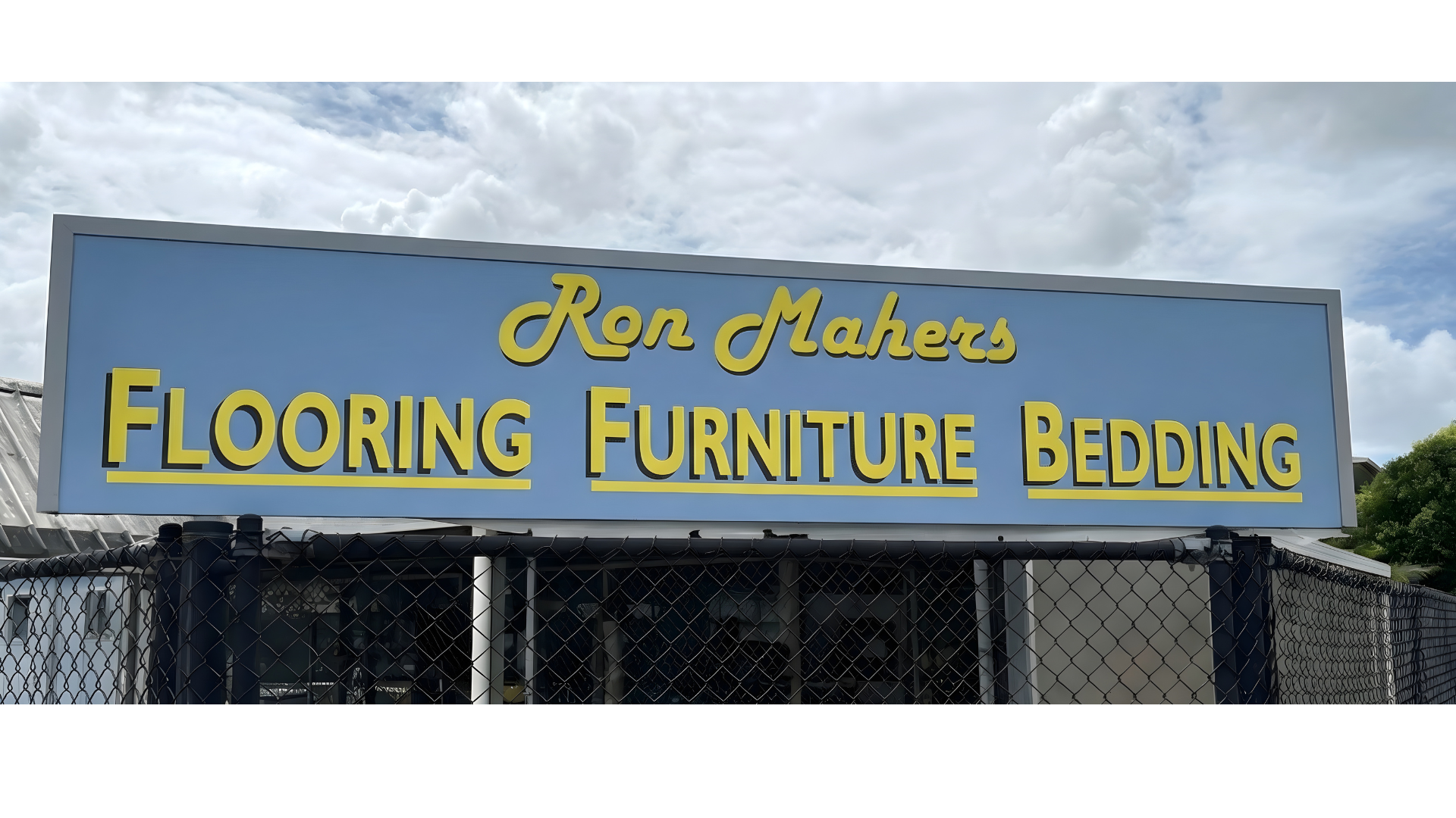 Storefront sign reads 'Ron Mahers Flooring Furniture Bedding' with a blue background and yellow text, under a cloudy sky.