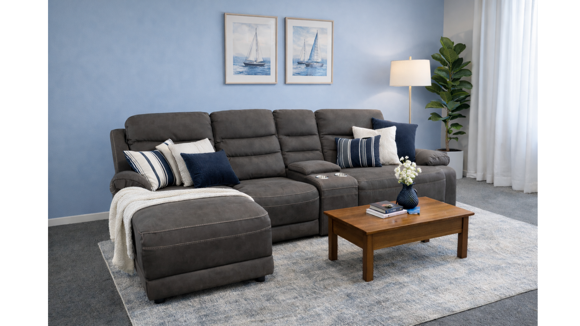 Living room with a dark gray sectional sofa, blue and white pillows, a wooden coffee table with books and a blue vase with white flowers, a tall lamp, and framed sailboat pictures on a blue wall.