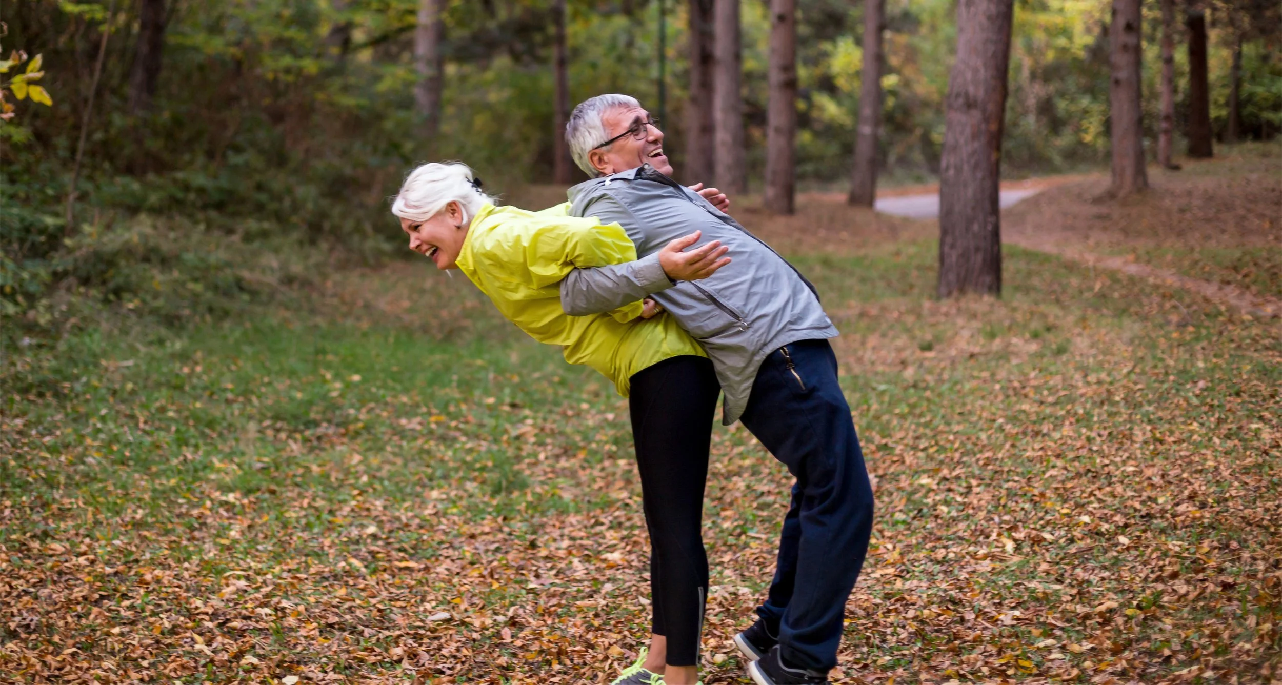 An elderly couple laughing and playing together in a wooded park during autumn, with the man holding the woman in a playful dip.