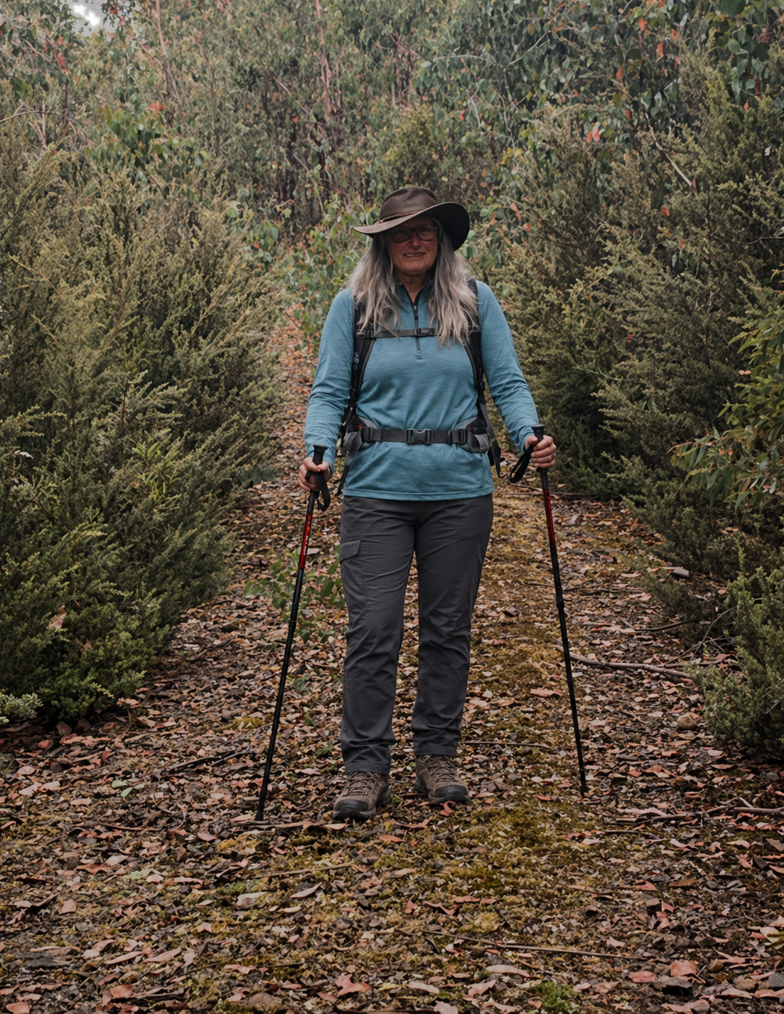 A woman hiking on a trail surrounded by green bushes, wearing a hat, glasses, a long-sleeve blue shirt, gray pants, and hiking boots, holding trekking poles.