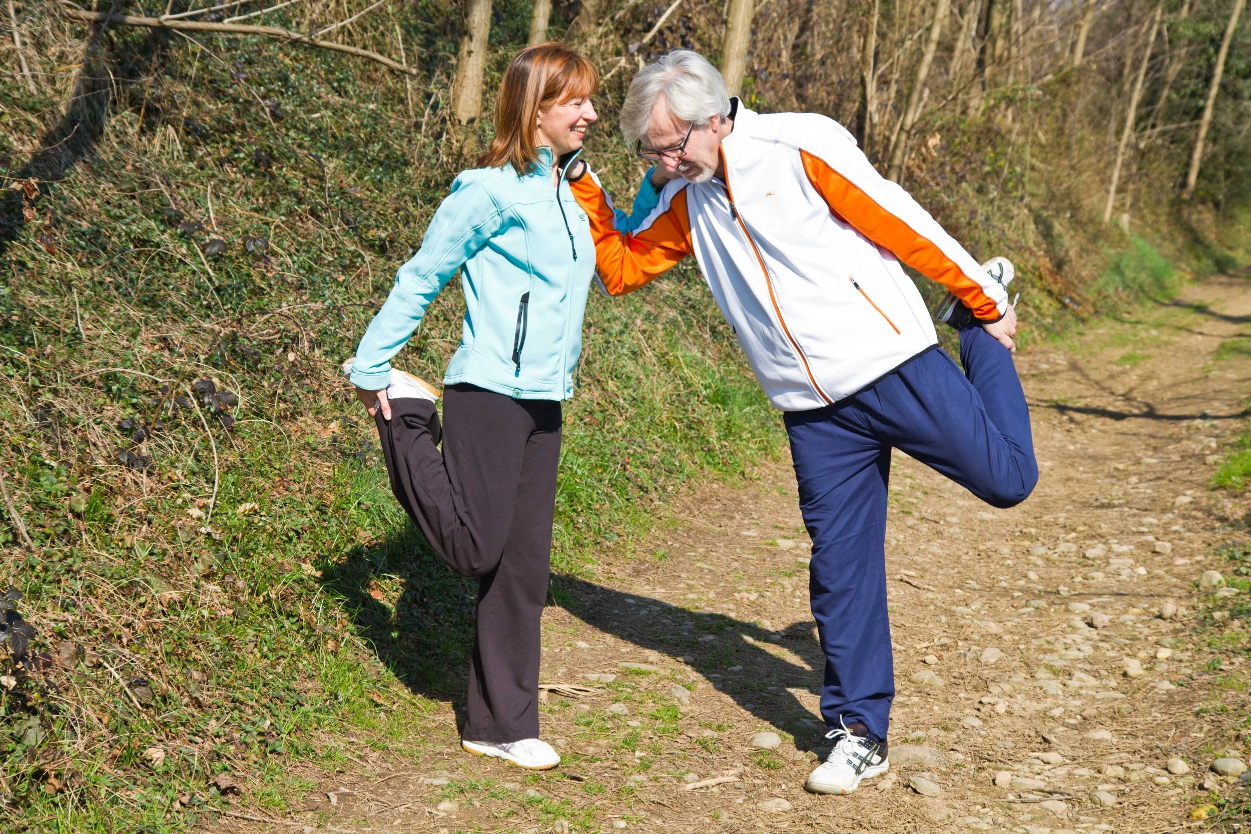 A man and woman on a dirt path in a forest, smiling and stretching together in exercise attire.