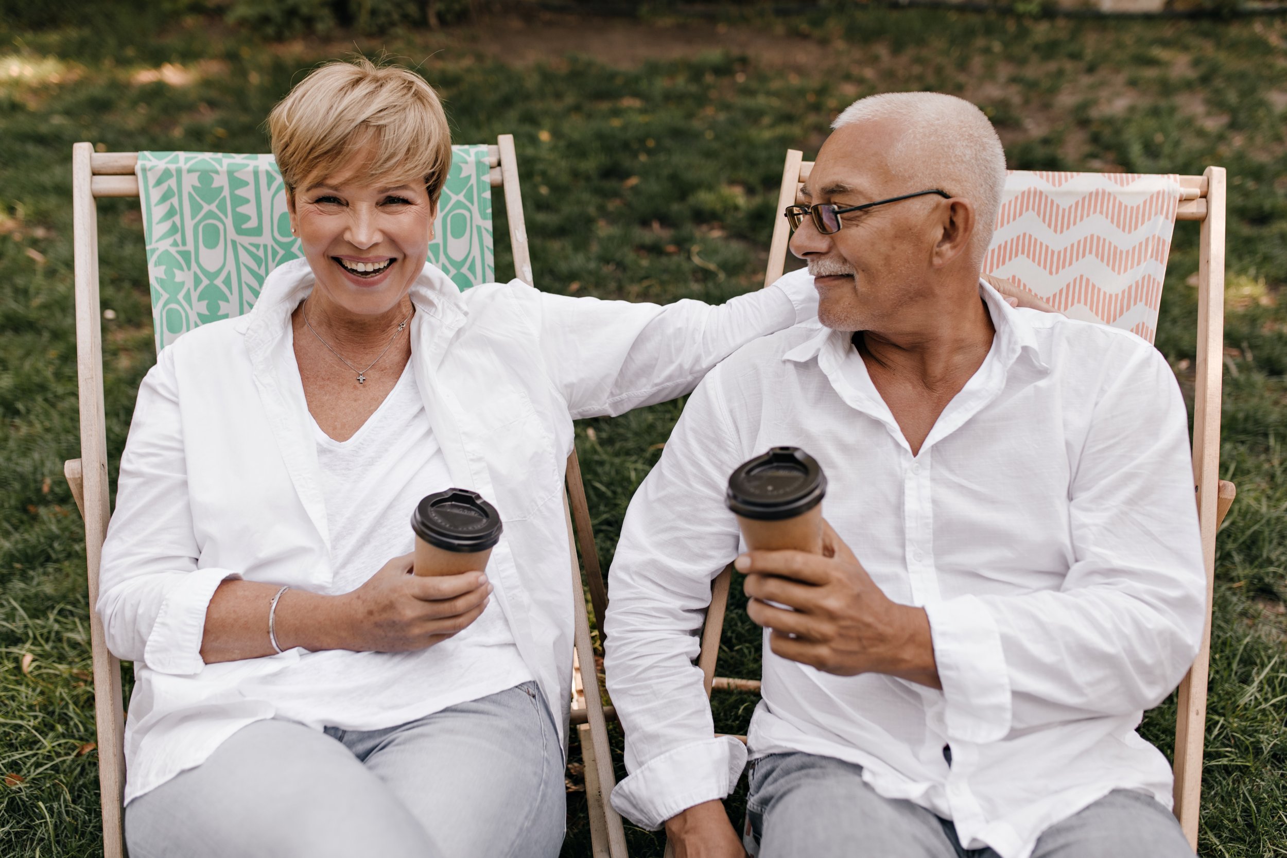 Smiling elderly woman and man sitting in deck chairs on grass, holding takeaway coffee cups, enjoying outdoor conversation.