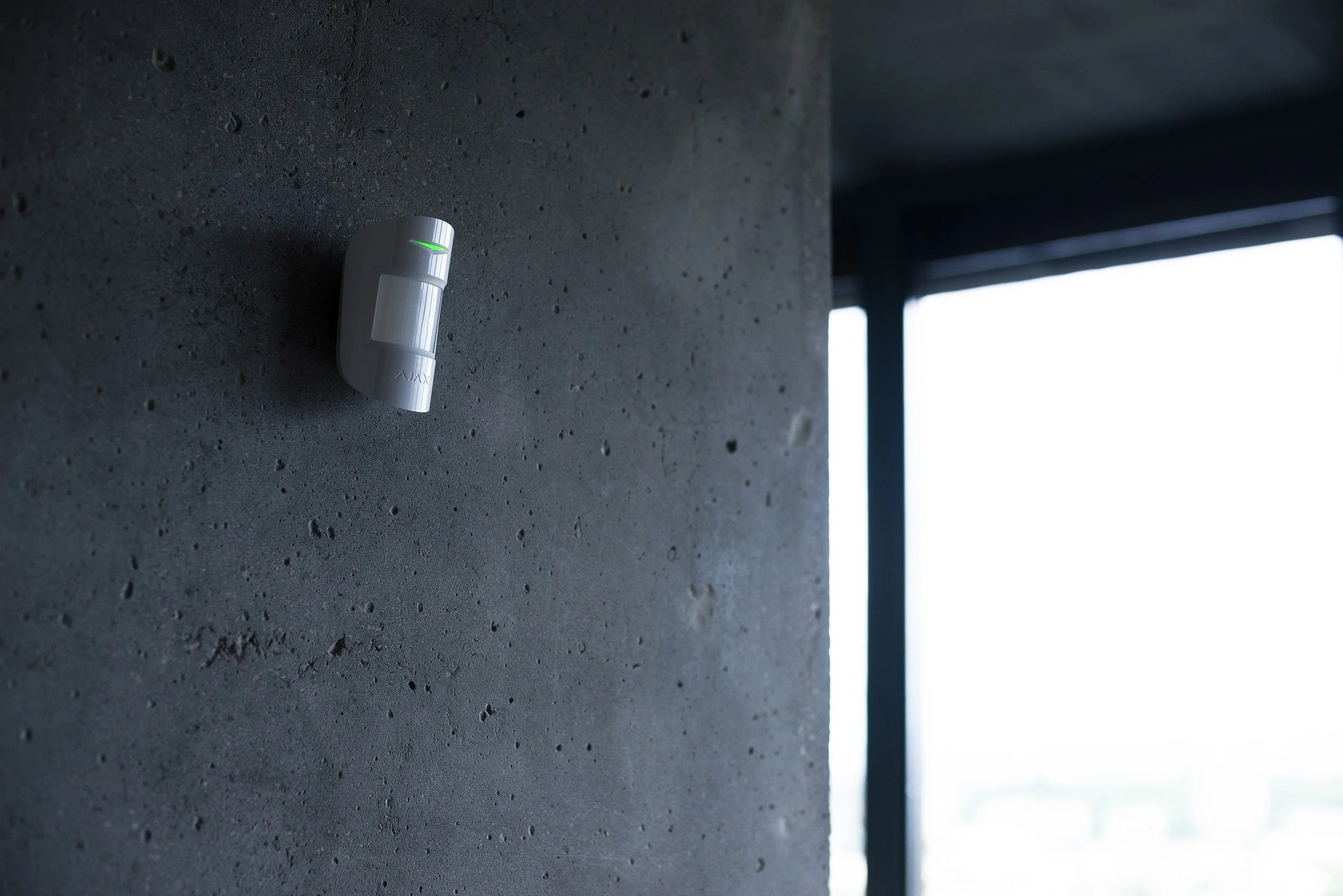 Capteur de mouvement blanc sur un mur en béton dans une pièce avec grande fenêtre laissant entrer de la lumière