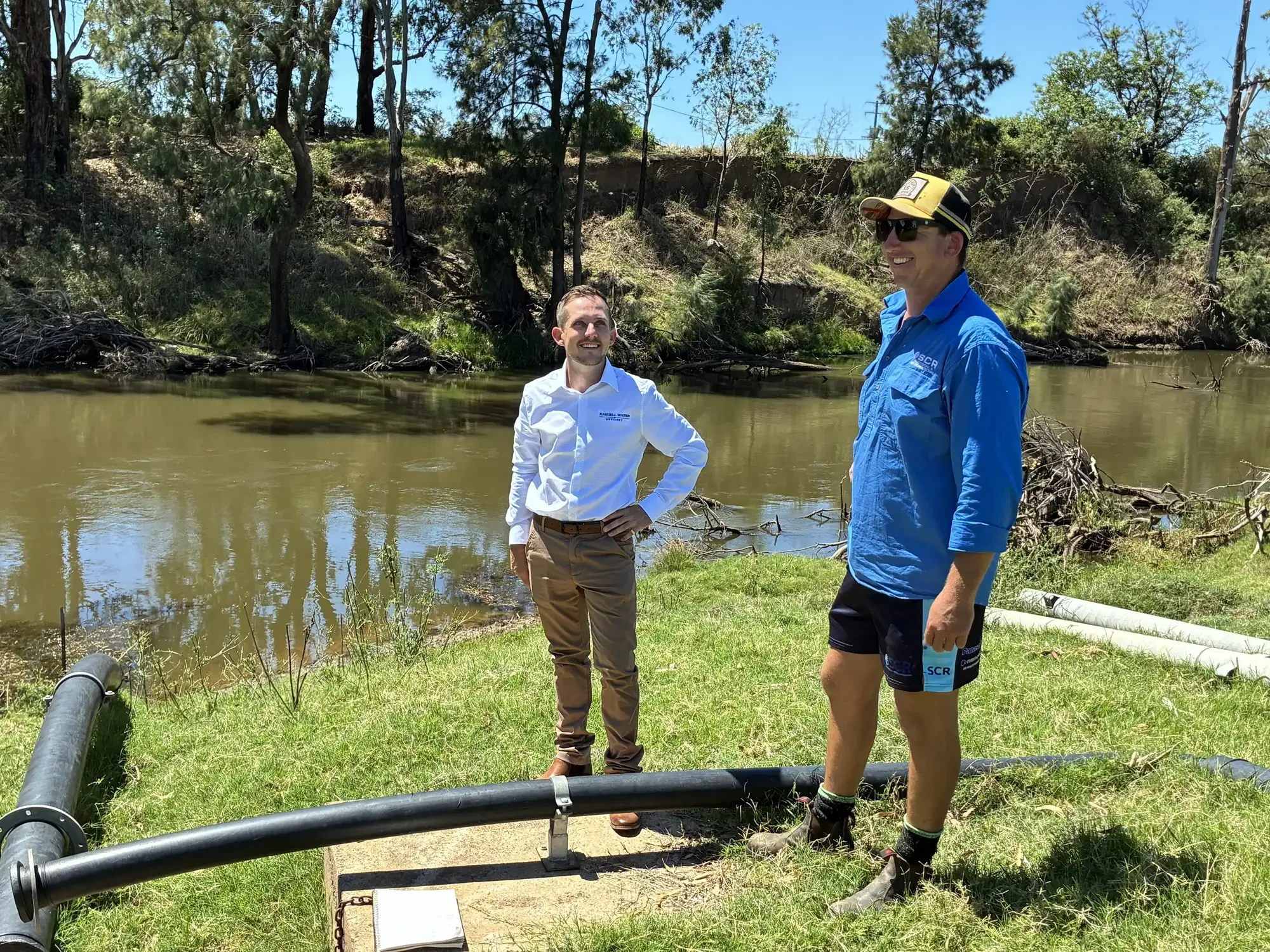 Rob standing with a client - they are smiling and appear to be engaged in a conversation. There is water equipment on the ground nearby, with trees and a grassy shoreline in the background.