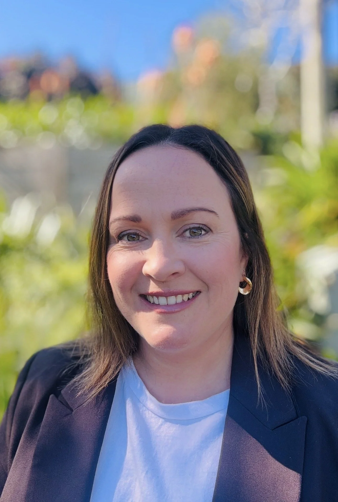 A woman with brown hair, green eyes, smiling, wearing a black blazer and a white shirt, standing outdoors on a sunny day with blurred trees and buildings in the background.
