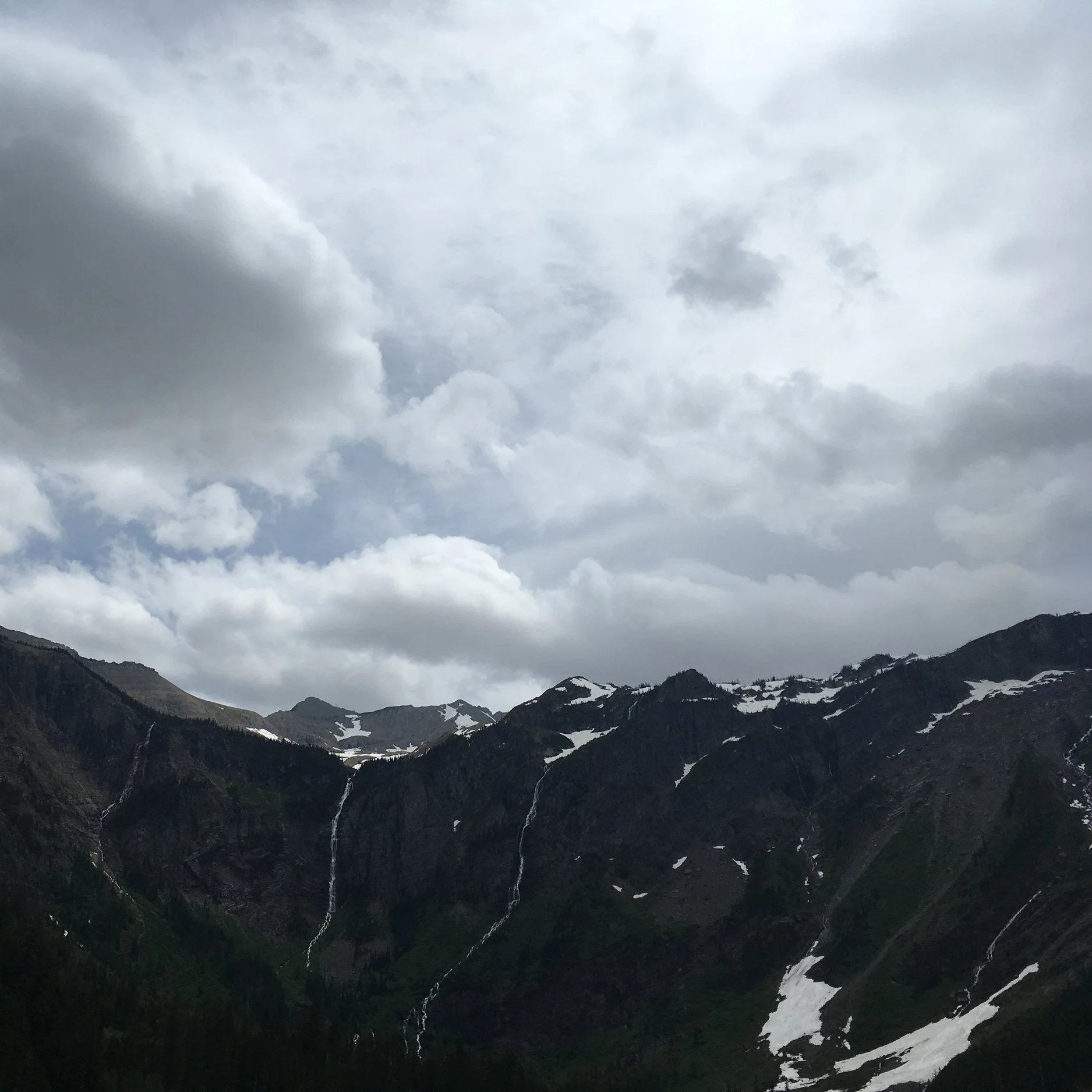 Mountain landscape with snow patches, waterfalls, and cloudy sky.