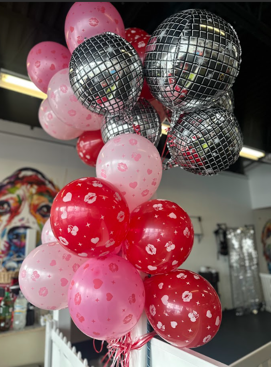 A cluster of pink, red, and silver balloons, including some metallic disco ball style balloons, in an indoor setting with colorful artwork in the background.
