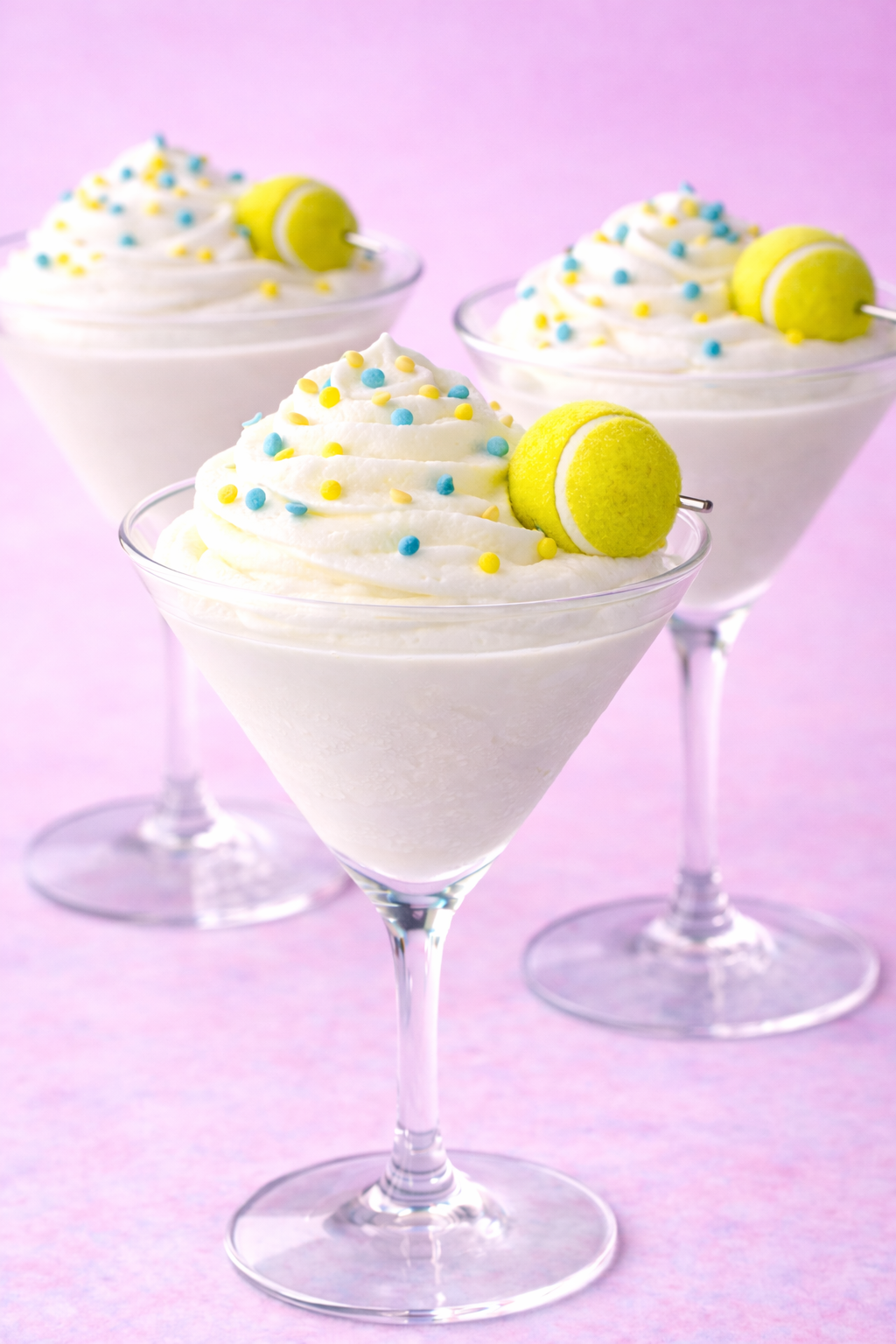 A whipped cream pupcup served in a martini glass, topped with a tennis ball garnish and blue and yellow sprinkles