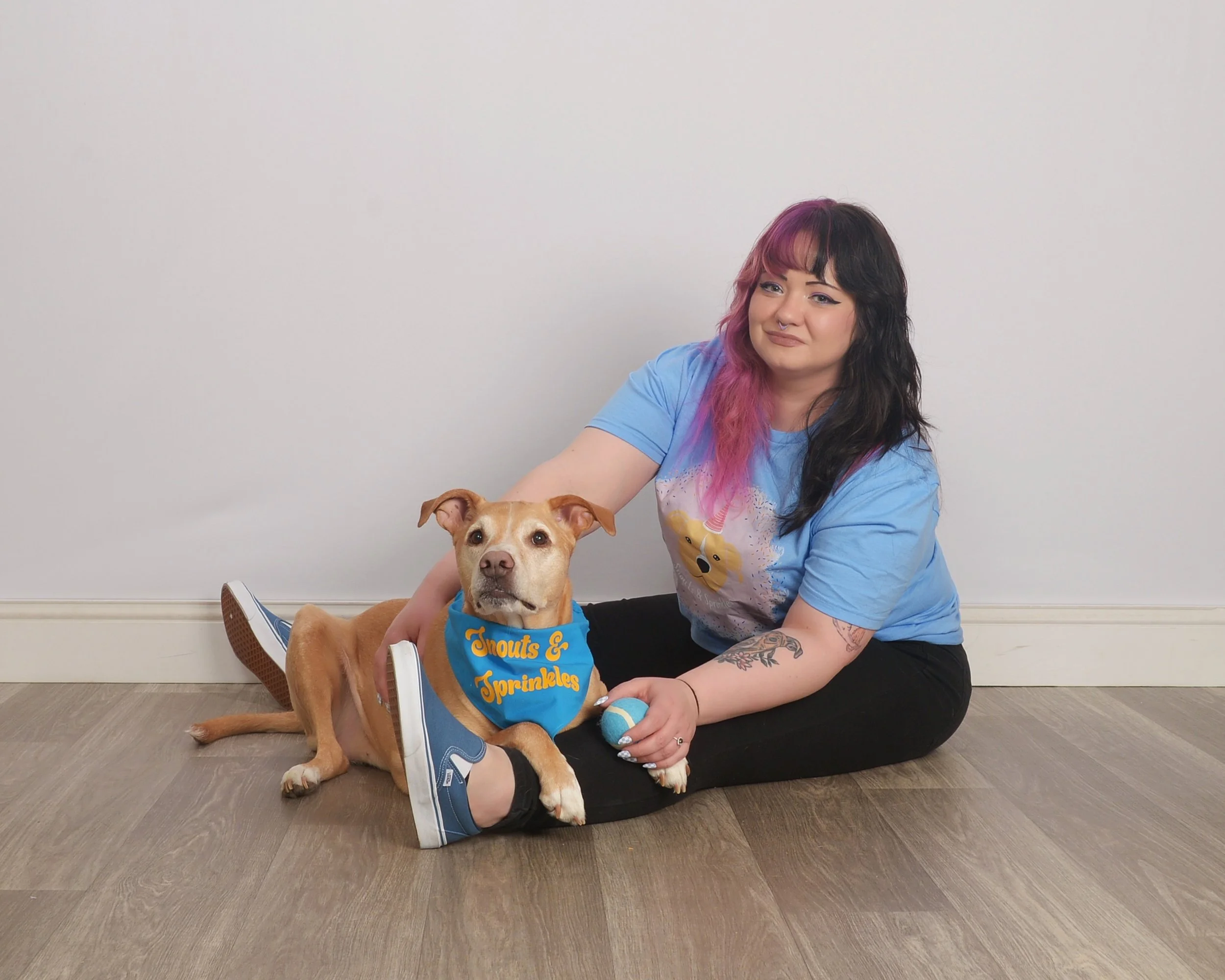 A woman with multicolored hair seated on the floor holding a tan dog wearing a bandana that says 'Knots & Sprinkles'. The woman is wearing a light blue T-shirt and black pants, and the dog is wearing sneakers.