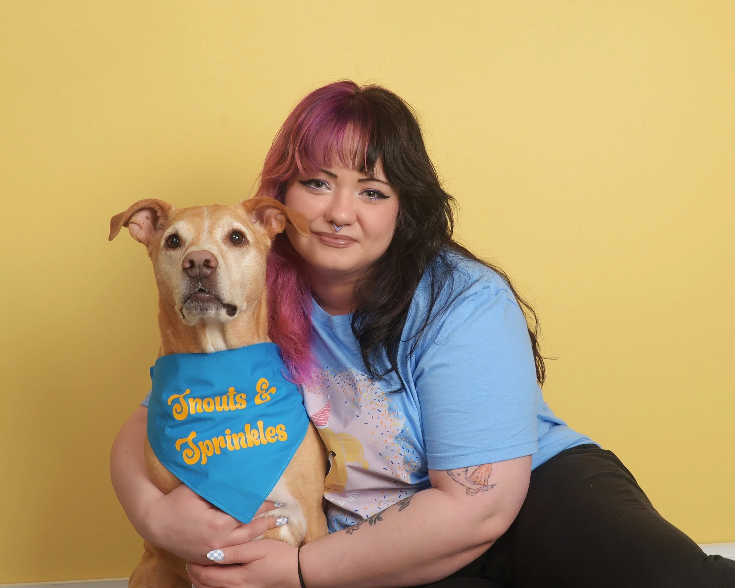 A woman with black and pink hair sitting on the floor, hugging a tan dog with a blue bandana that says 'Knots & Sprinkles', against a yellow background.