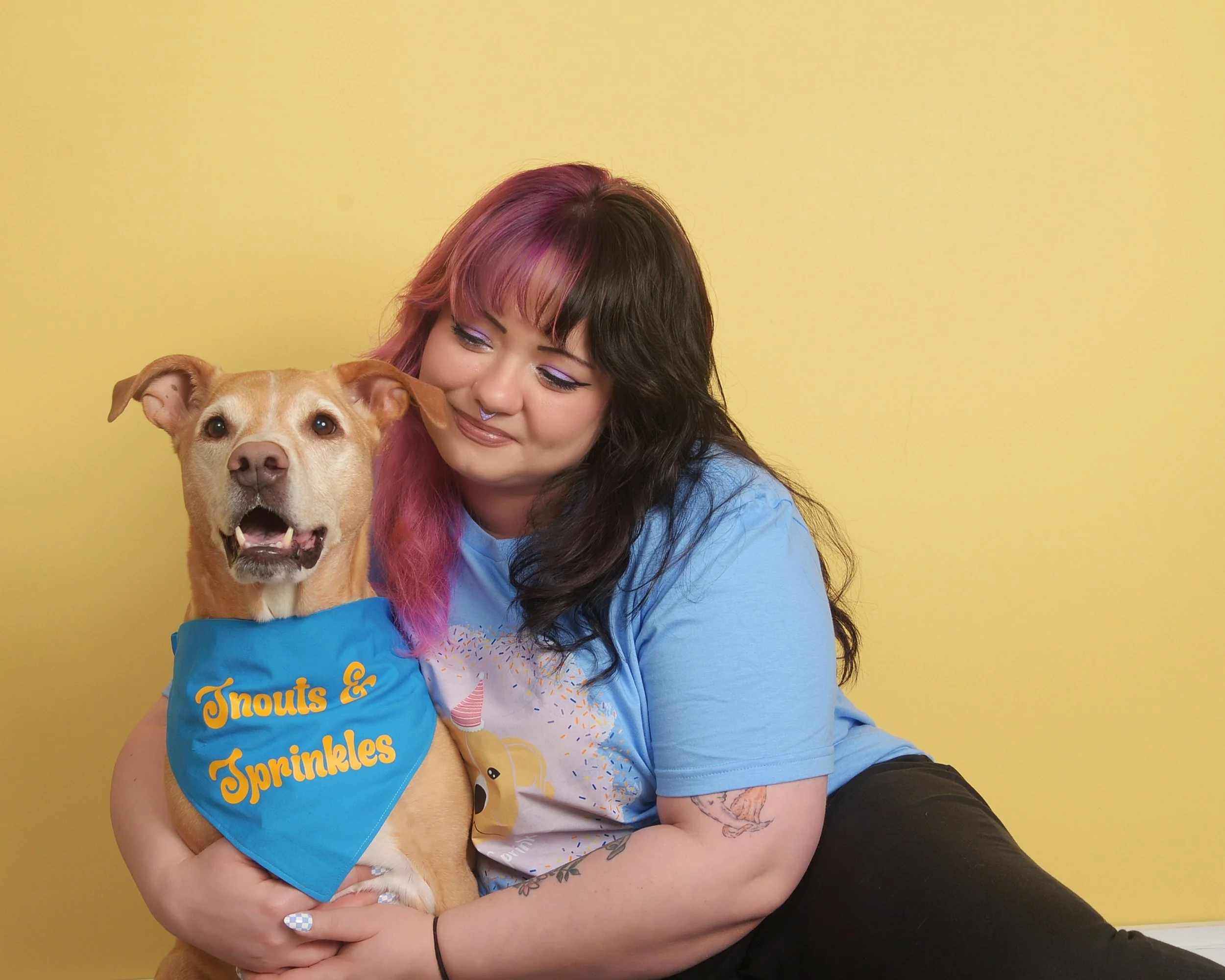 A woman with colorful hair and tattoos hugging a happy dog wearing a blue bandana with yellow text that says 'Inouts & Sprinkles' against a yellow background.