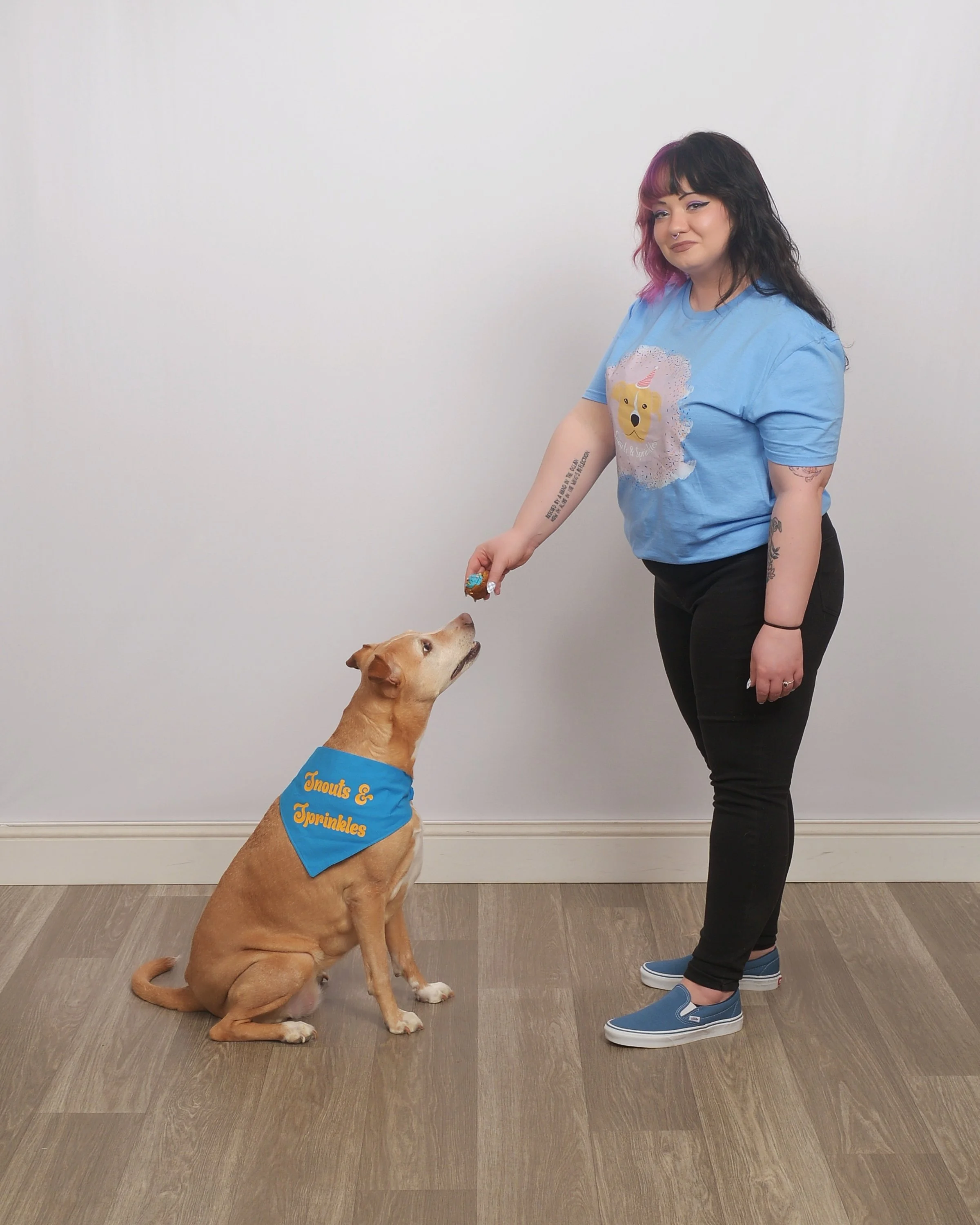 A woman with black and pink hair wearing a blue shirt with a unicorn on it, black pants, and blue slip-on shoes, is holding a treat in her hand. The dog, wearing a blue bandana with yellow text that reads "Knouts & Sprinkles," is sitting on the floor and looking up at the woman, reaching for the treat.