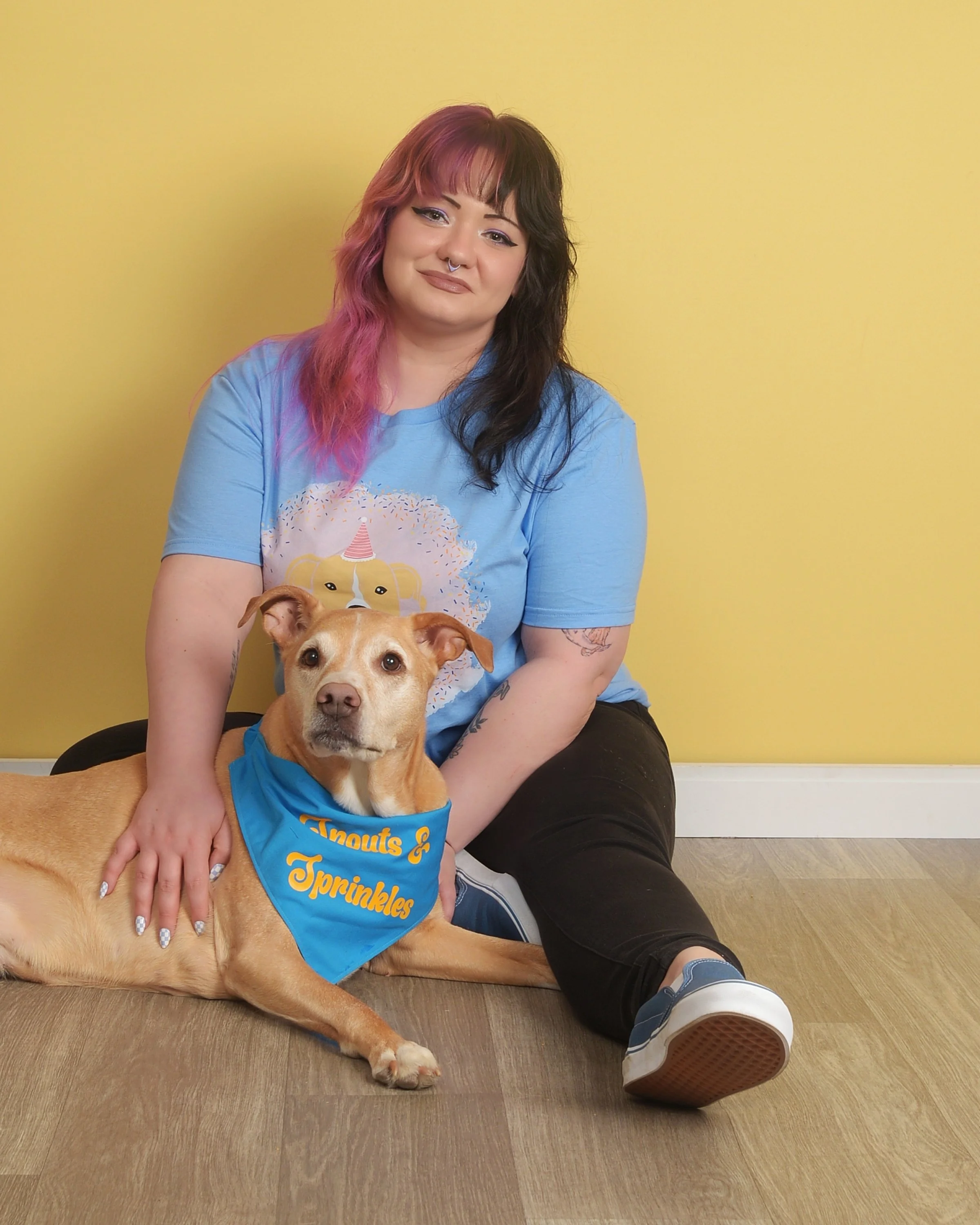 A woman with pink and black hair sitting on the floor with a tan dog wearing a blue bandana that reads 'Knouts & Sprinkles' against a yellow wall background.