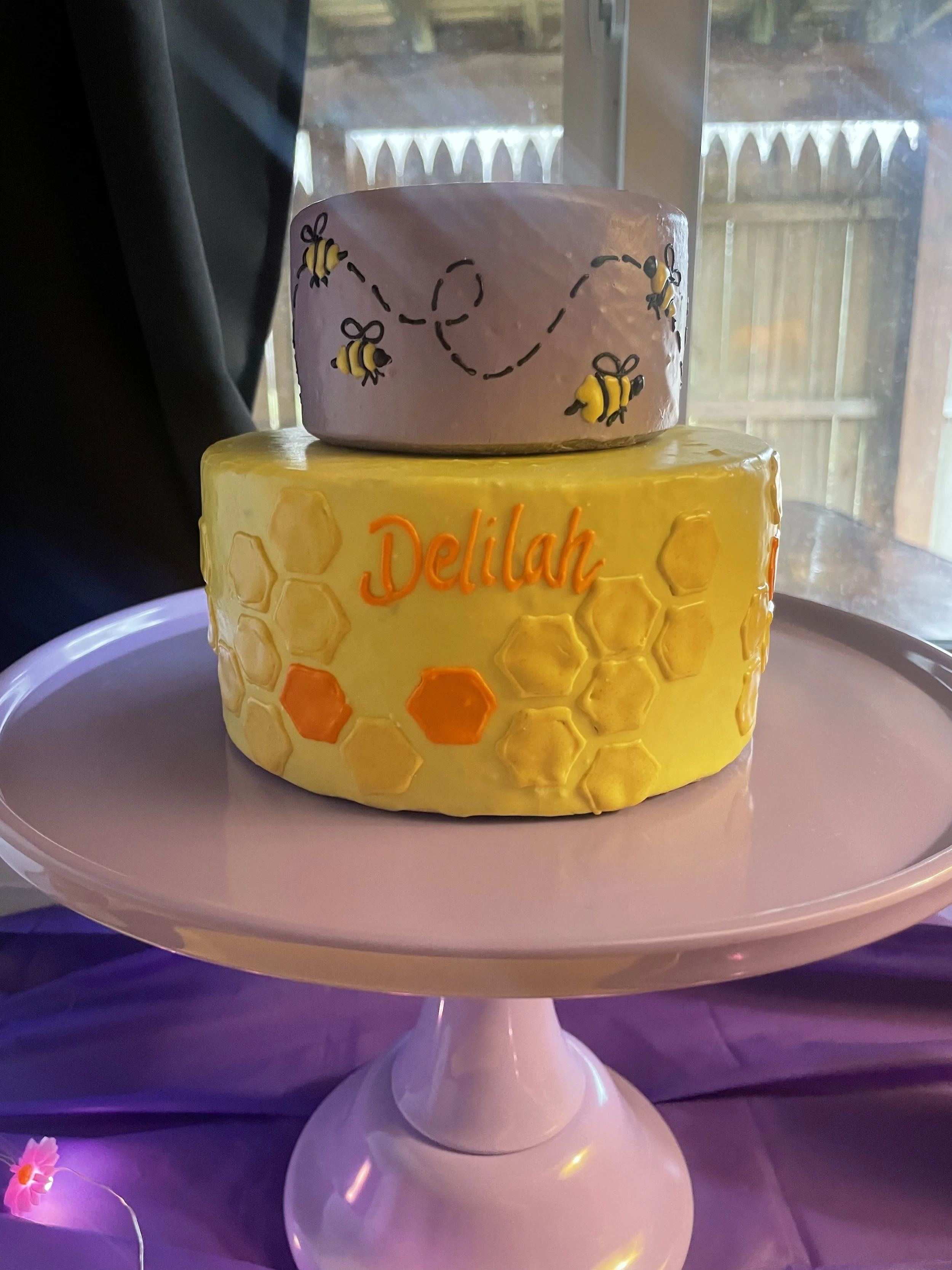 Two decorated cakes on a white cake stand, one with a honeycomb pattern and the word 'Delilah,' and the other with bee and flower illustrations, and a dashed heart-shaped trail, in front of a window with a garden view.