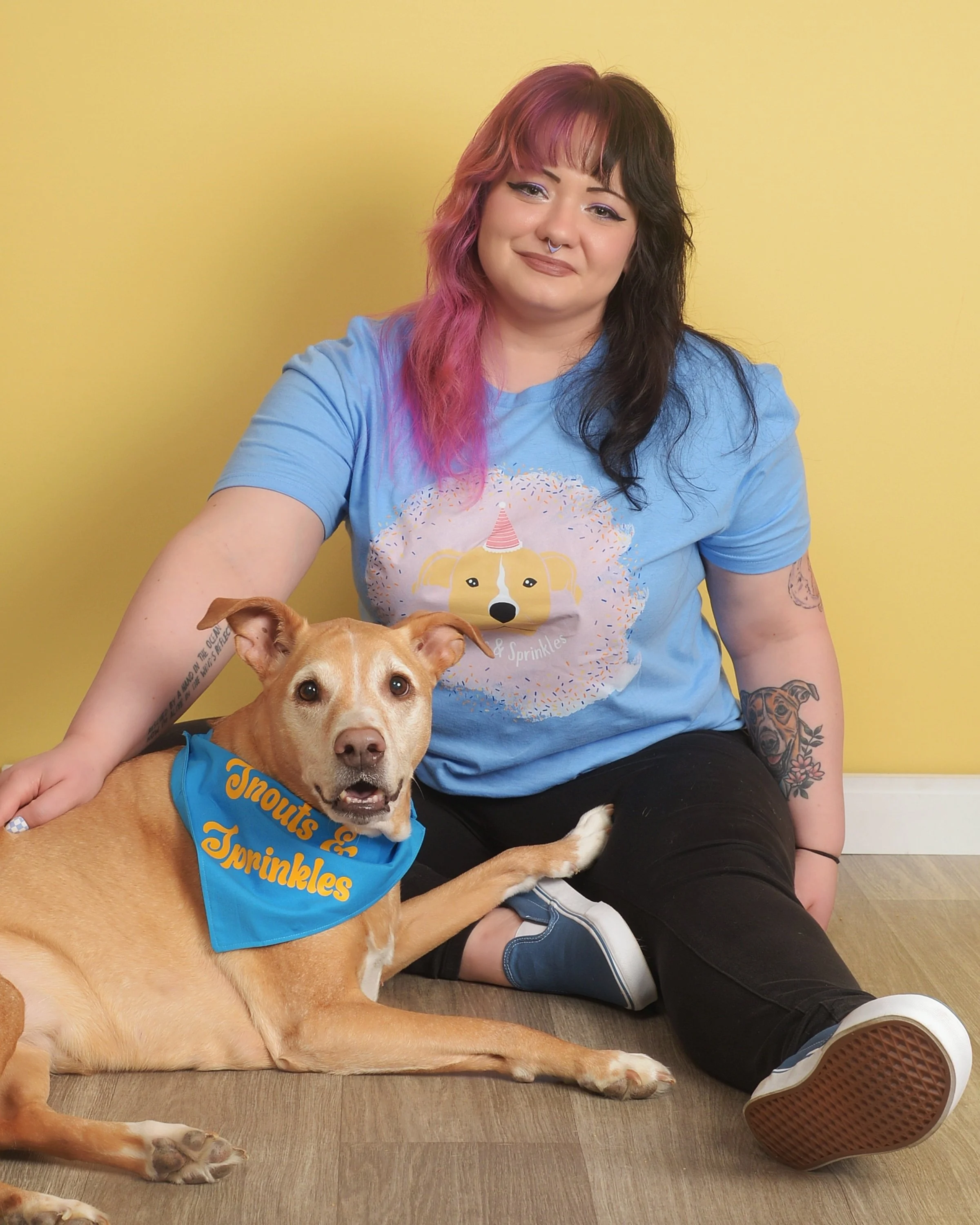 A woman with pink and black hair sitting on the floor next to a tan dog with a blue bandana reading "Snouts & Sprinkles". The woman is wearing a blue T-shirt with a dog illustration and black pants, with tattoos on her arms. They are against a yellow